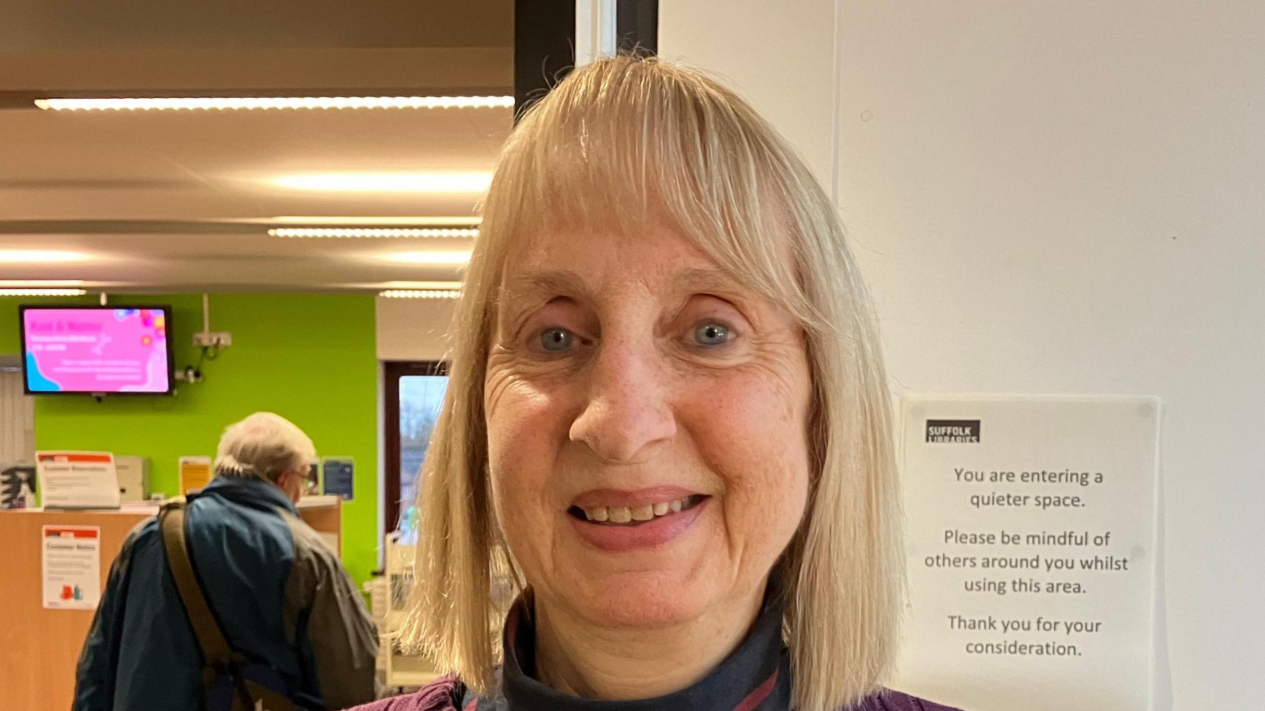 Elizabeth McAlpine smiles at the camera. She has straight, blonde hair that sits just above her shoulders. A man can be seen in the background with his back turned away from the camera. 