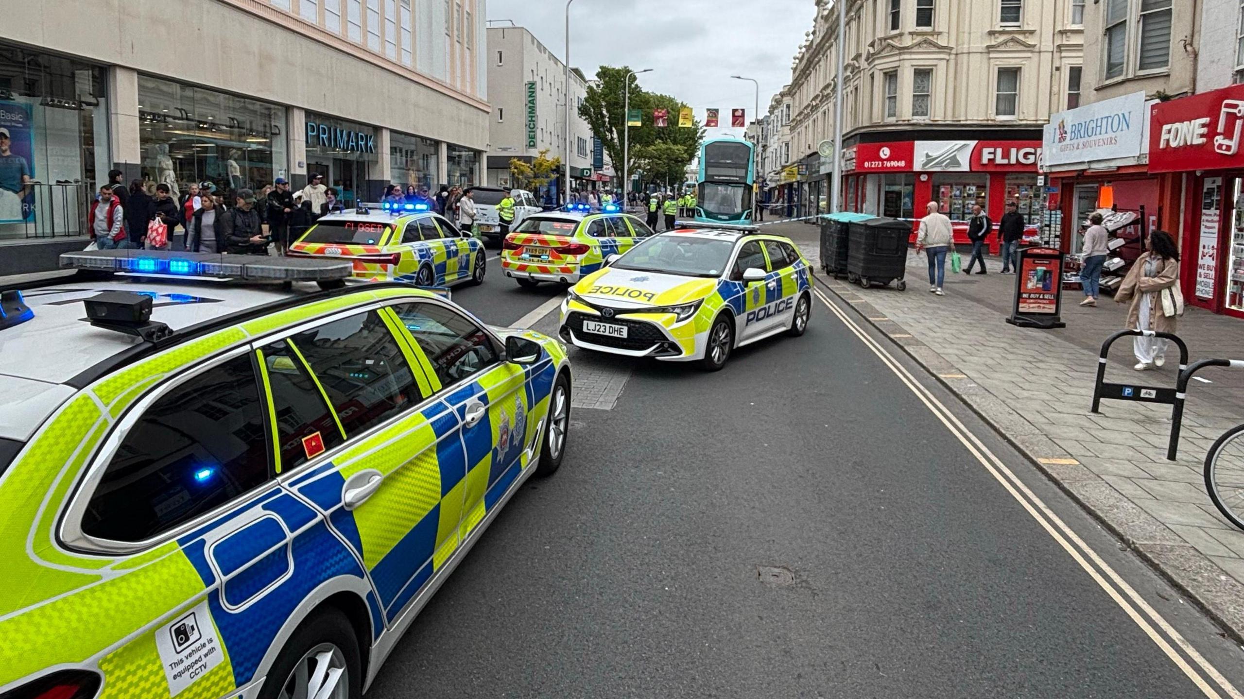 Four police cars parked on a road in Brighton, in front of a cordon of police tape. A double decker bus is behind the cordon.