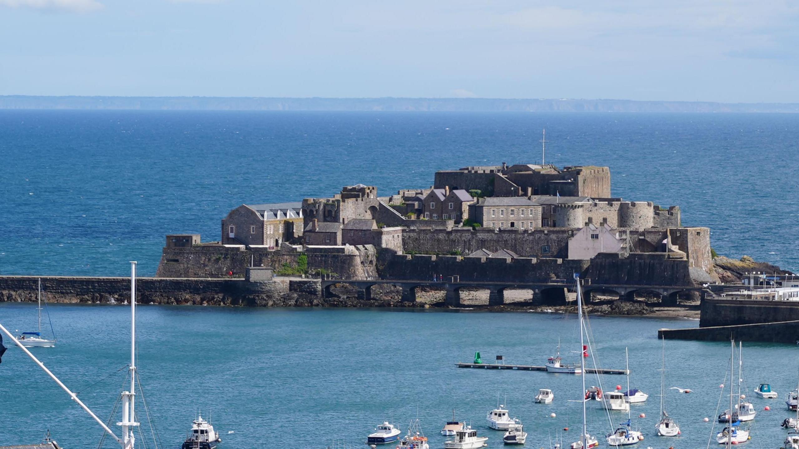 Castle Cornet from afar. A large fort structure standing inbetween St Peter Port Harbour and the open sea. 

The castle itself is brown stone and has a number of different levels, with walls that have inlets for gun emplacements. 