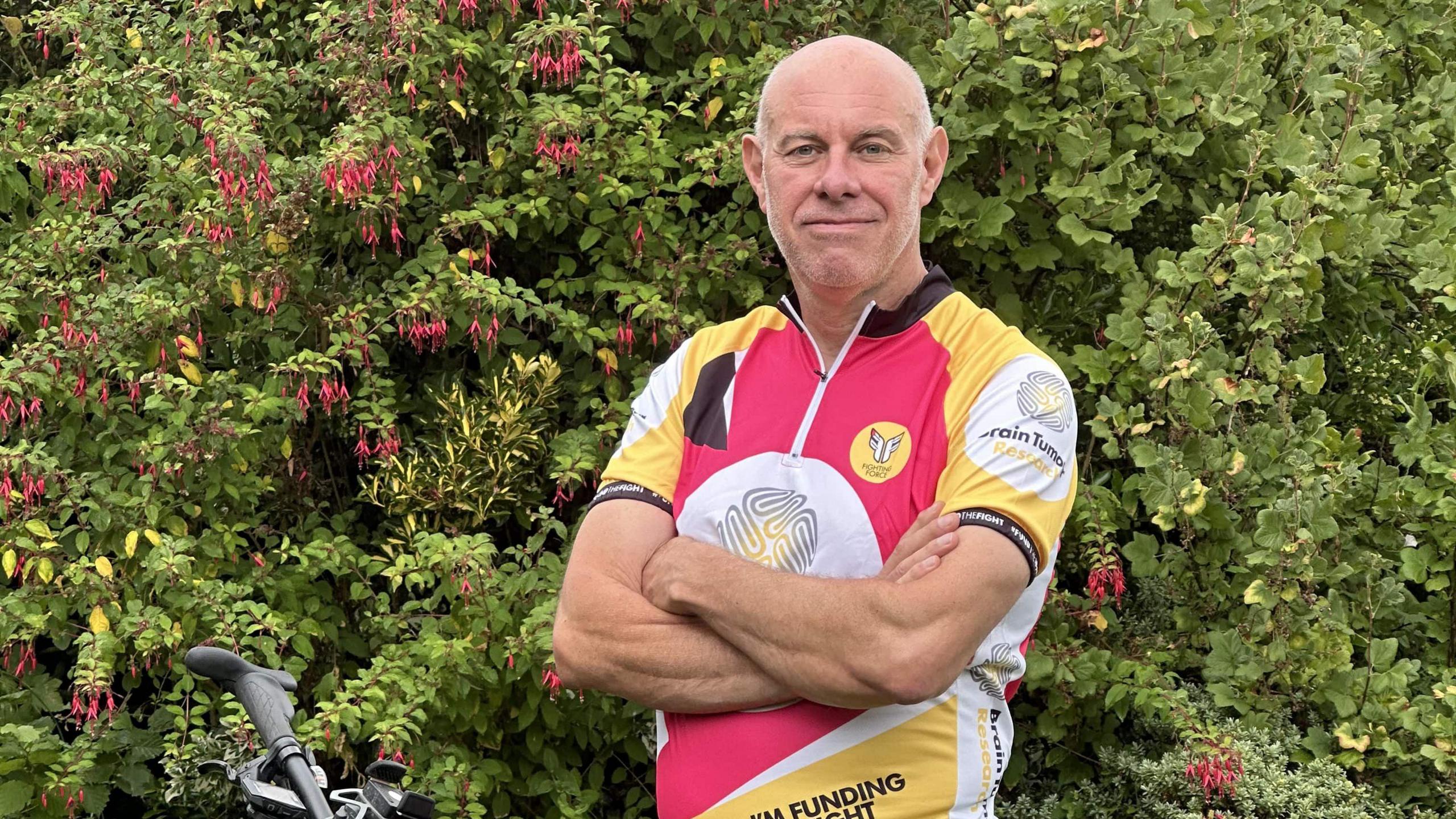 John Starns wearing a pink, yellow and white cycle top looking to camera in front of a hedge
