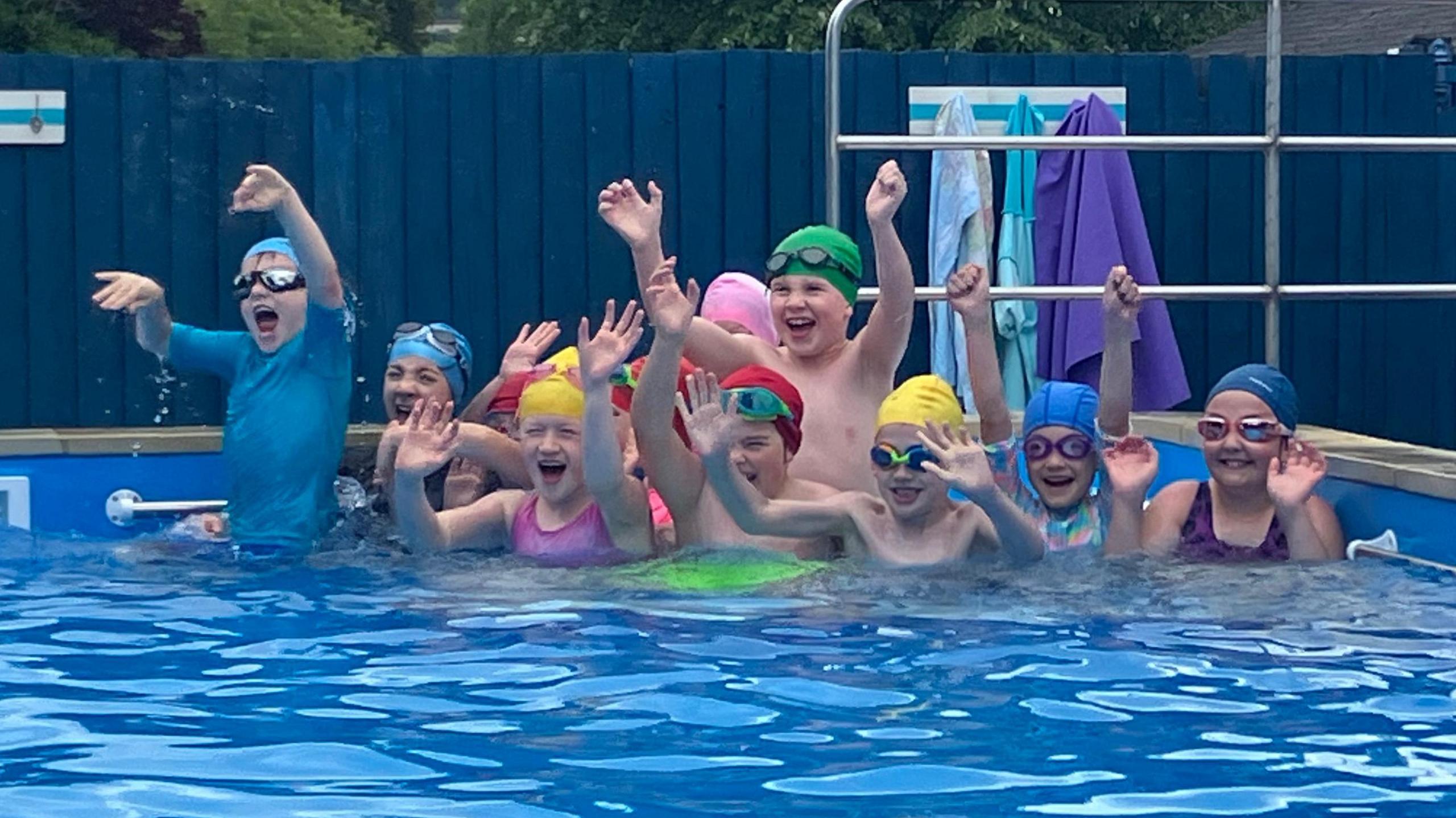A photo showing a group of children in a swimming pool wearing swimming hats and goggles and they are waving