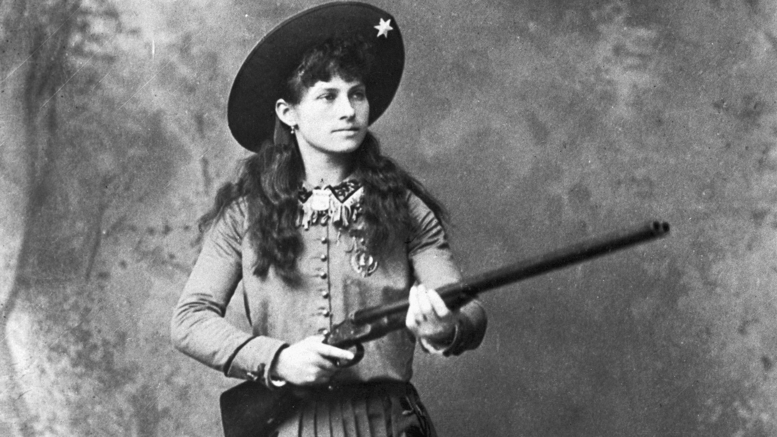 A black and white photo of a young Annie Oakley wearing wild west outfit with hat and holding a rifle.
