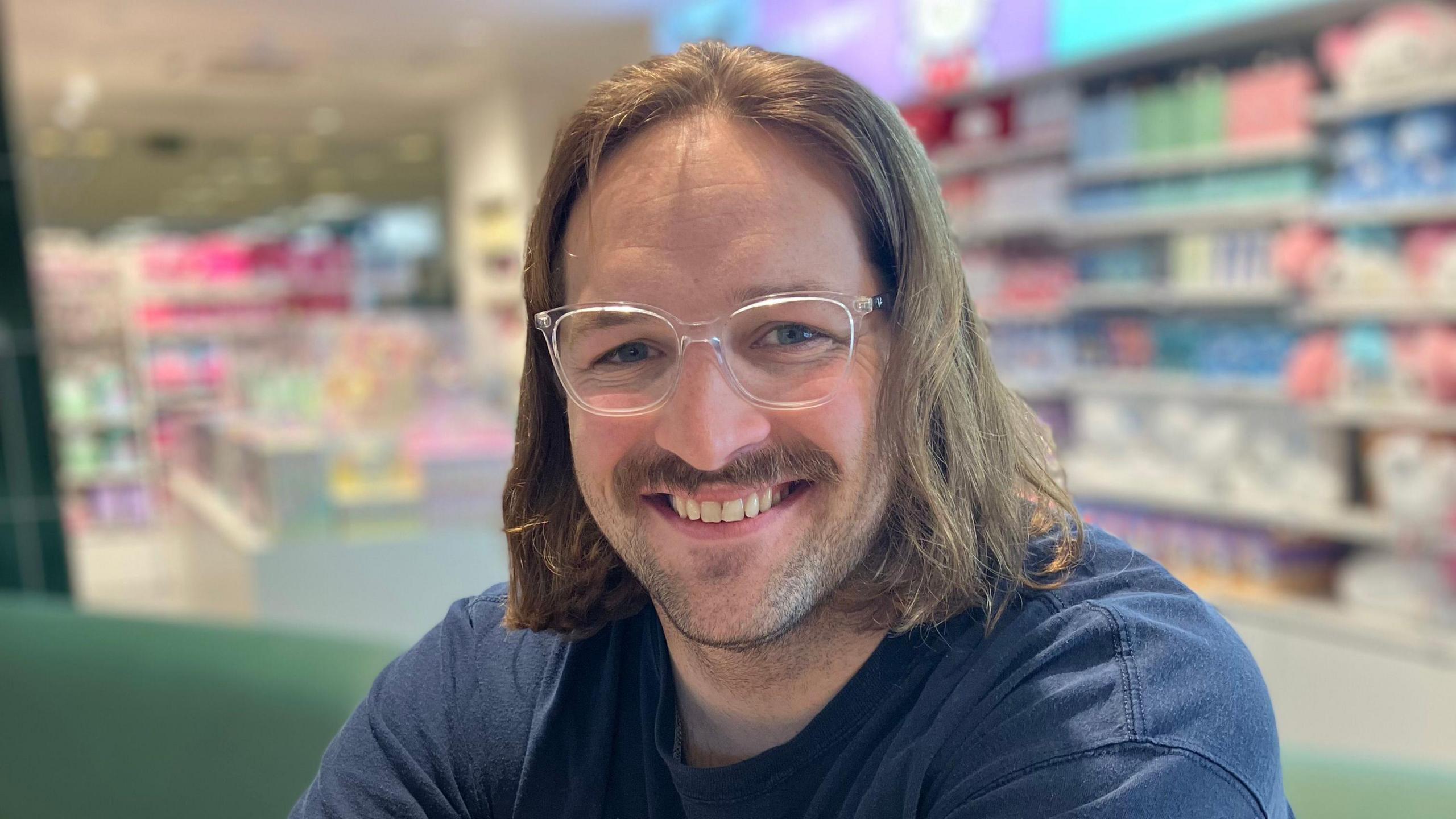Daniel is smiling at the camera sitting down on a green sofa. Behind him is toys that are blurred out. He has clear glasses and blue eyes and shoulder length dark blond hair in a curtain parting. His tshirt is dark blue.