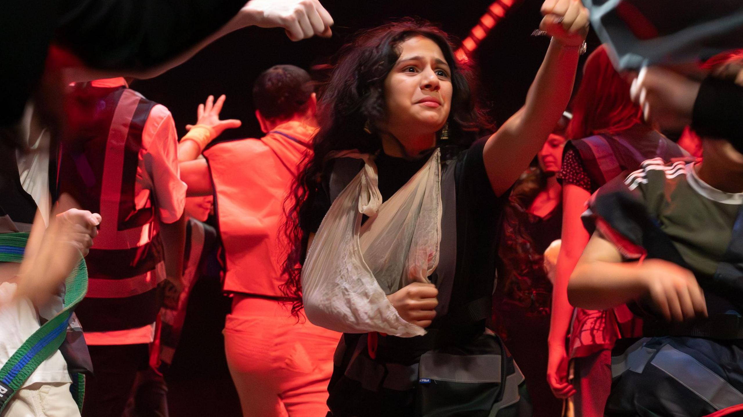 A teenage girl with her arm in a sling raises her fist looking anguished, while other children dance around her