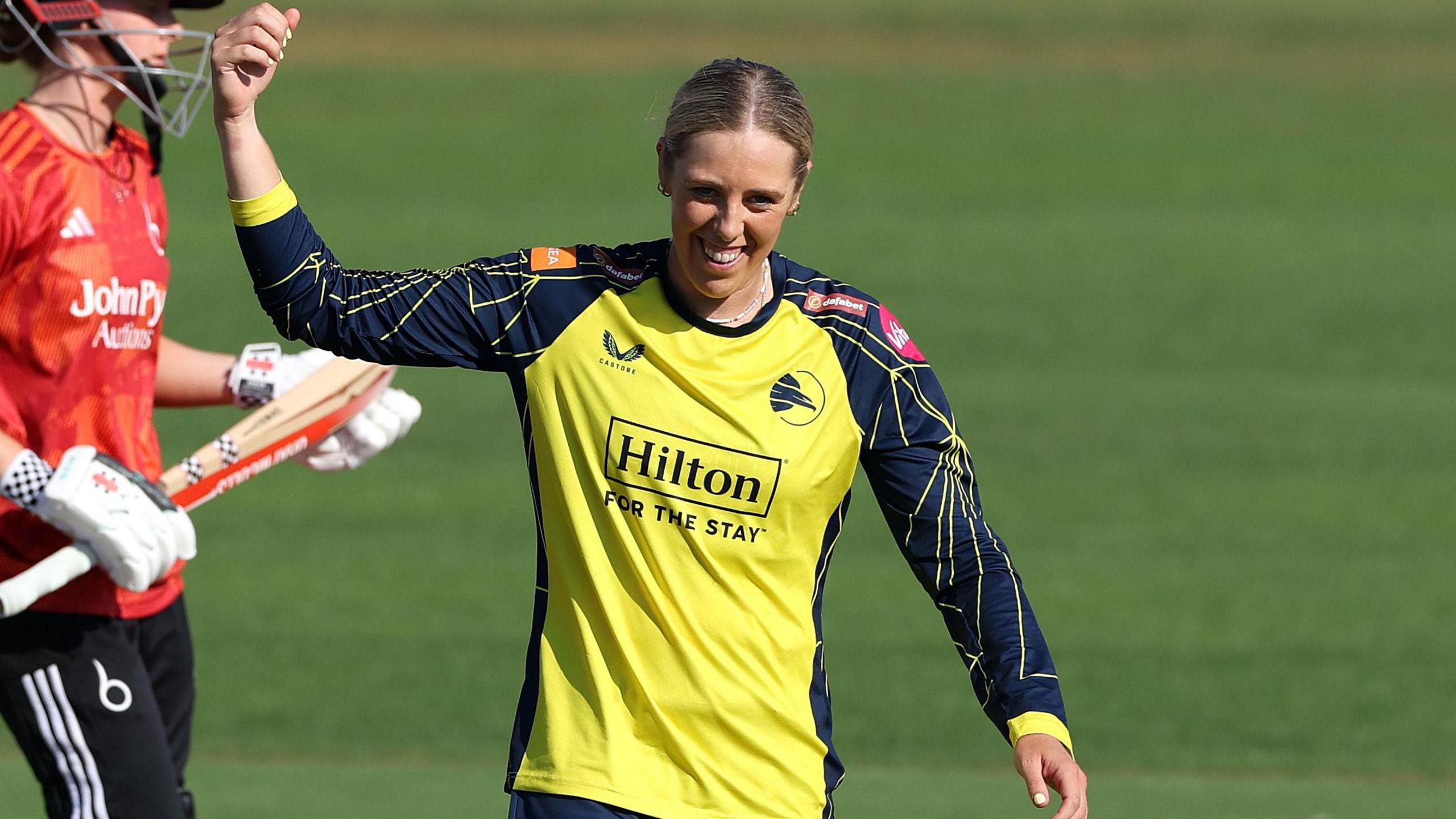 Georgia Adams, wearing Hampshire's yellow shirt with navy blue sleeves and collar, celebrates taking a wicket by raising her right hand while smiling