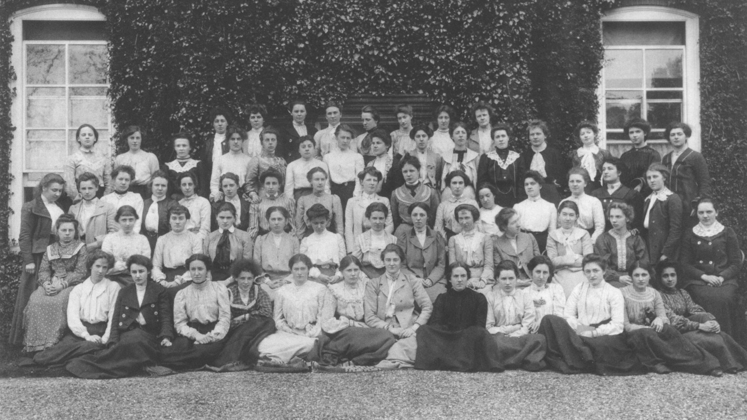 A group image in black and white from 1902 showing female students at Newnham College. They are arranged in a group shot in five rows. They are wearing long dresses and all have their hair up in different hairdos of the time. They are standing in front of a building covered in vegetation.