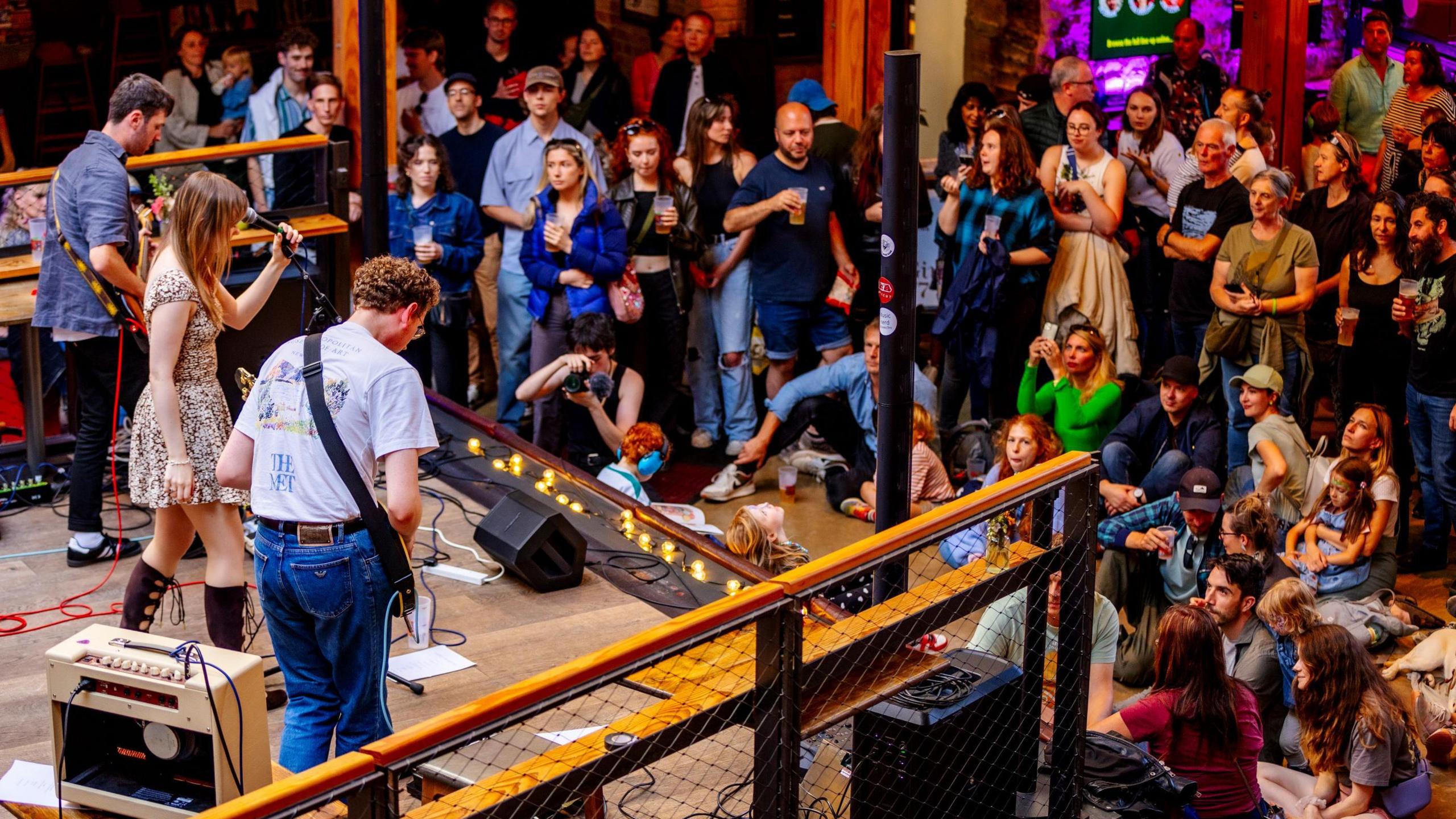 Two male musicians and a female singer perform for a crowd at Old City Sounds in the centre of Bristol. Some of the crowd are seated and others are taking pictures or dancing. The musicians are on a low platform