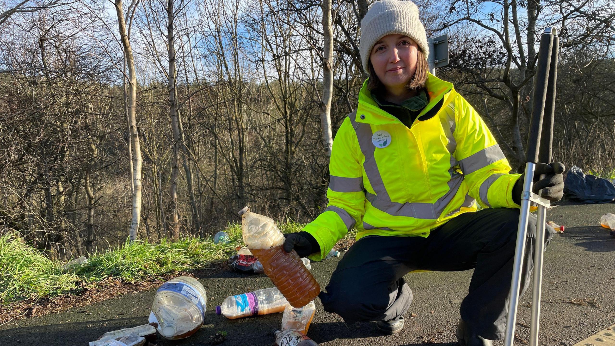 A woman in a hi-vis jacket, with litter picking equipment, is crouched in a lay-by holding a large plastic bottle, with other litter in the foreground and background