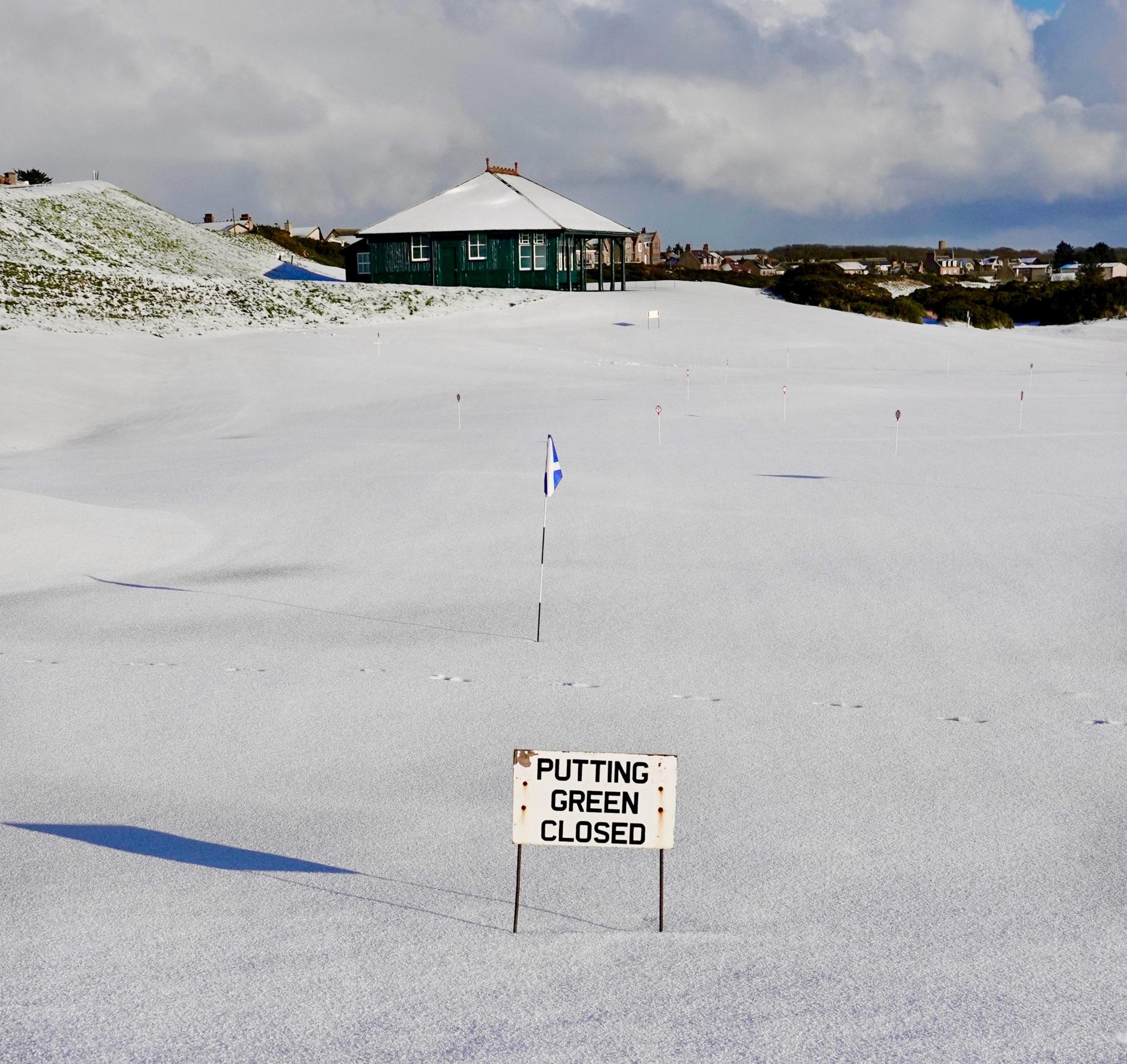 A golf course with flags covered in snow and a sign saying "putting green closed"