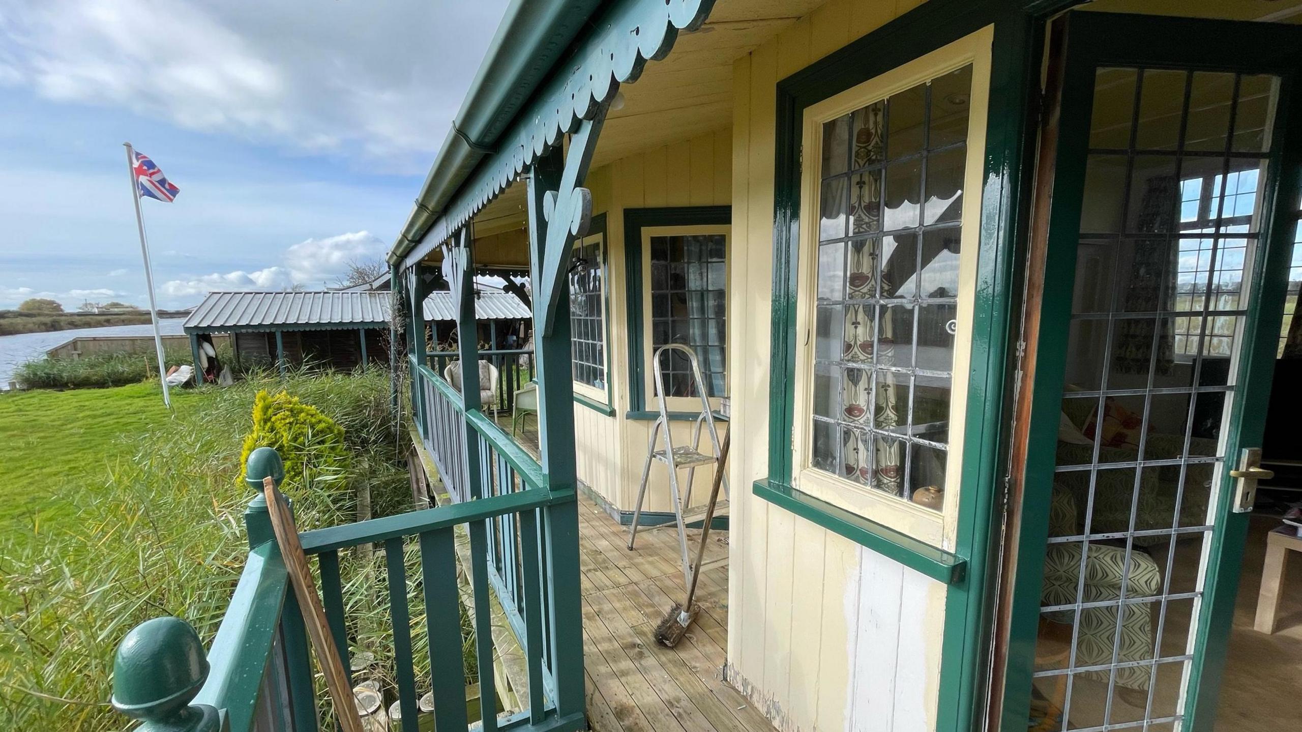 The view from Towerview's veranda, which is painted green wood. The walls of the house are cream painted wood and it has a wooden floor. Beyond the house is grass, with a flagpole flying a Union Flag, and beyond that can be seen the Broads waterway