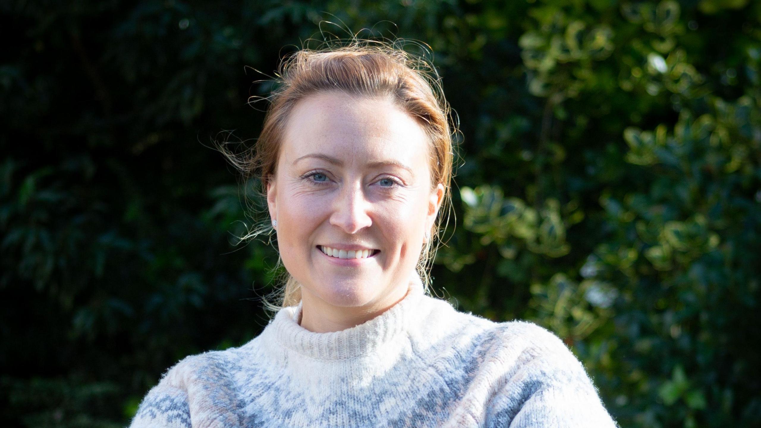 Lillie Pusey smiling at the camera wearing a blue and white jumper, with green foliage behind her. 