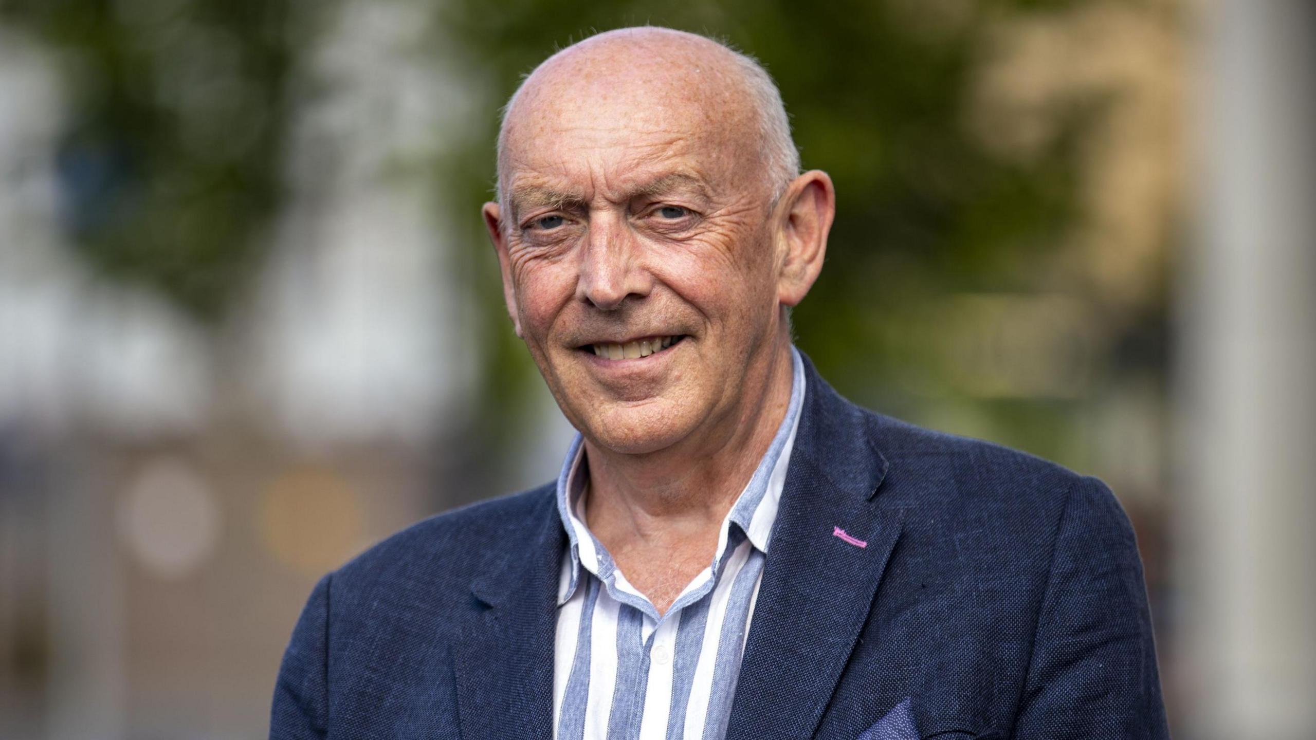 Peter Sheridan looking at the camera wearing a dark suit jacket and a open-neck striped shirt