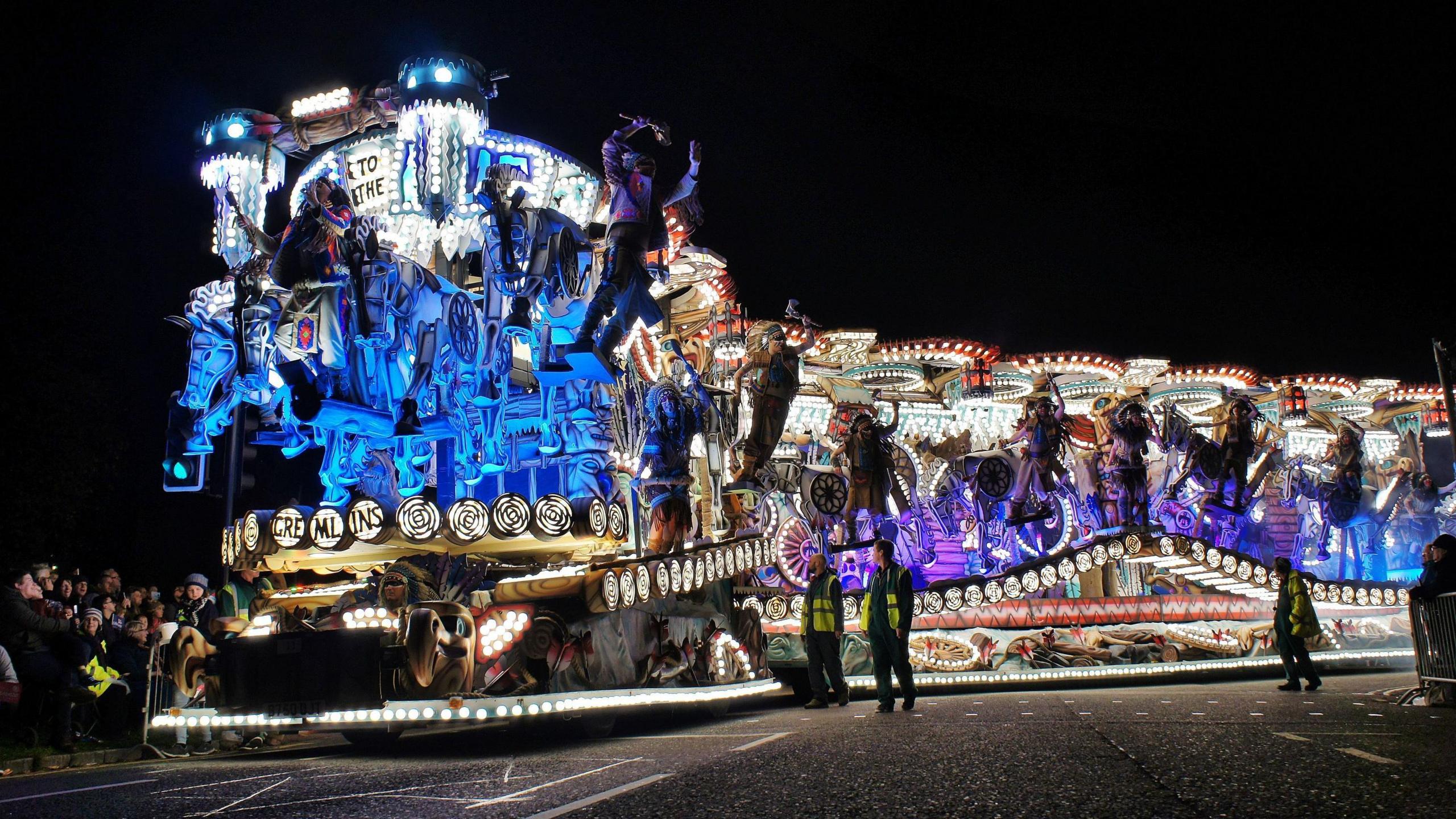 A long, brightly-lit carnival cart with people hanging off it all the way down its length. It is photographed from the side.