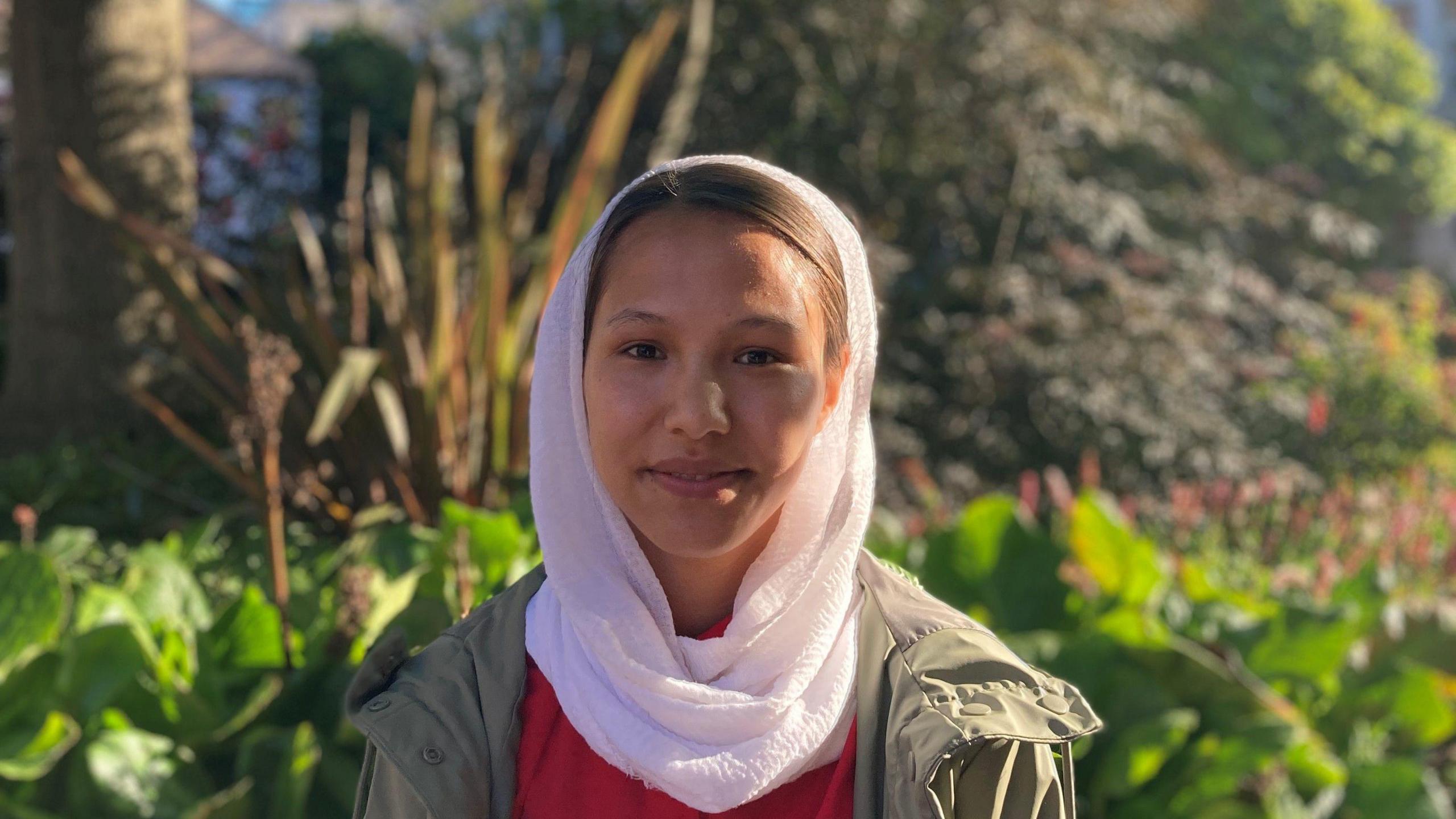 Zarifa Asghari stands in front of some trees. She is facing the camera and smiling. She is wearing a jacket and a white Hijab.