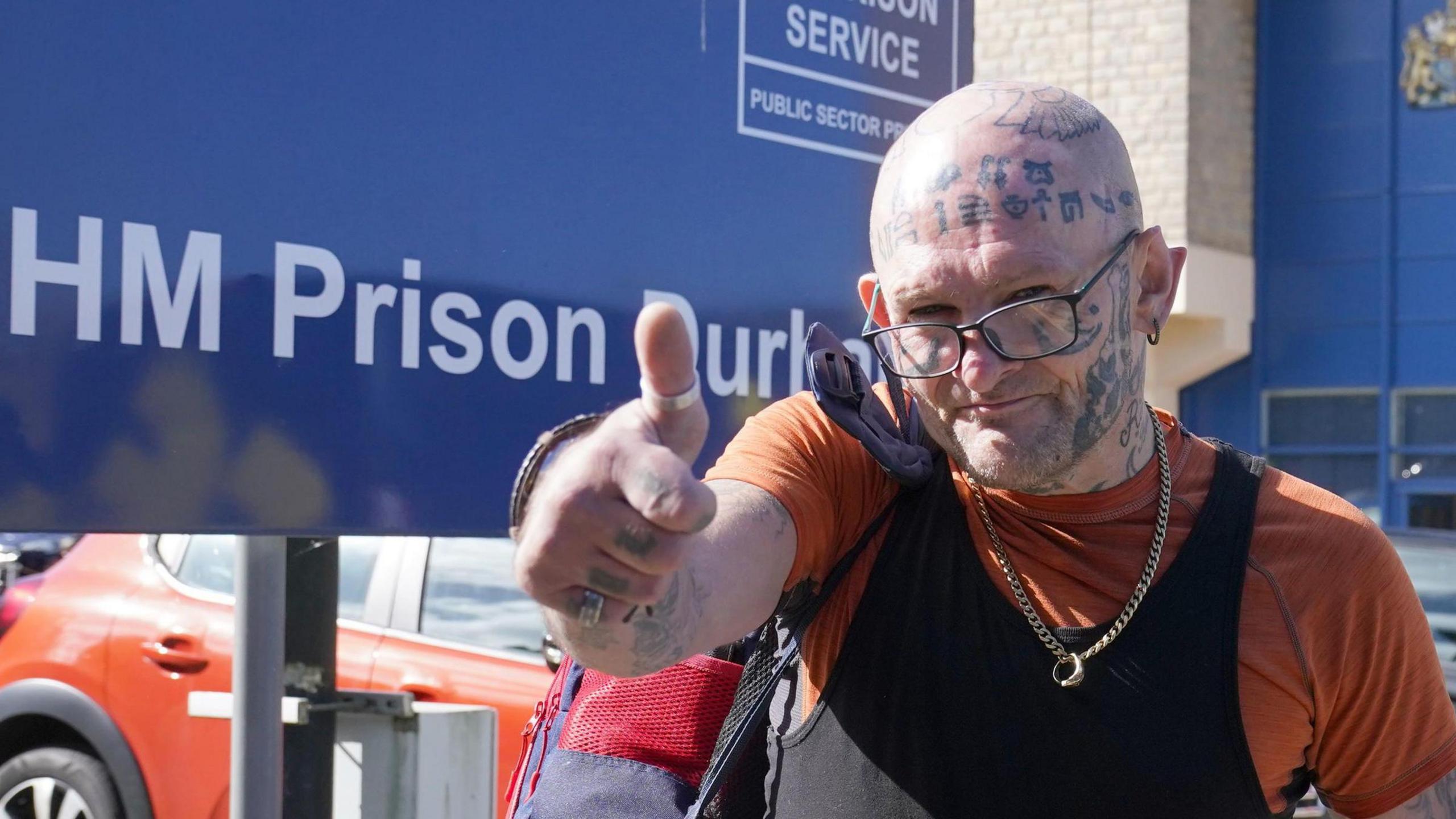 A photo of Jason Hoganson standing in front of a sign for HM Prison Durham with his thumb up. He is bald with a tattooed hand, arm and face. 