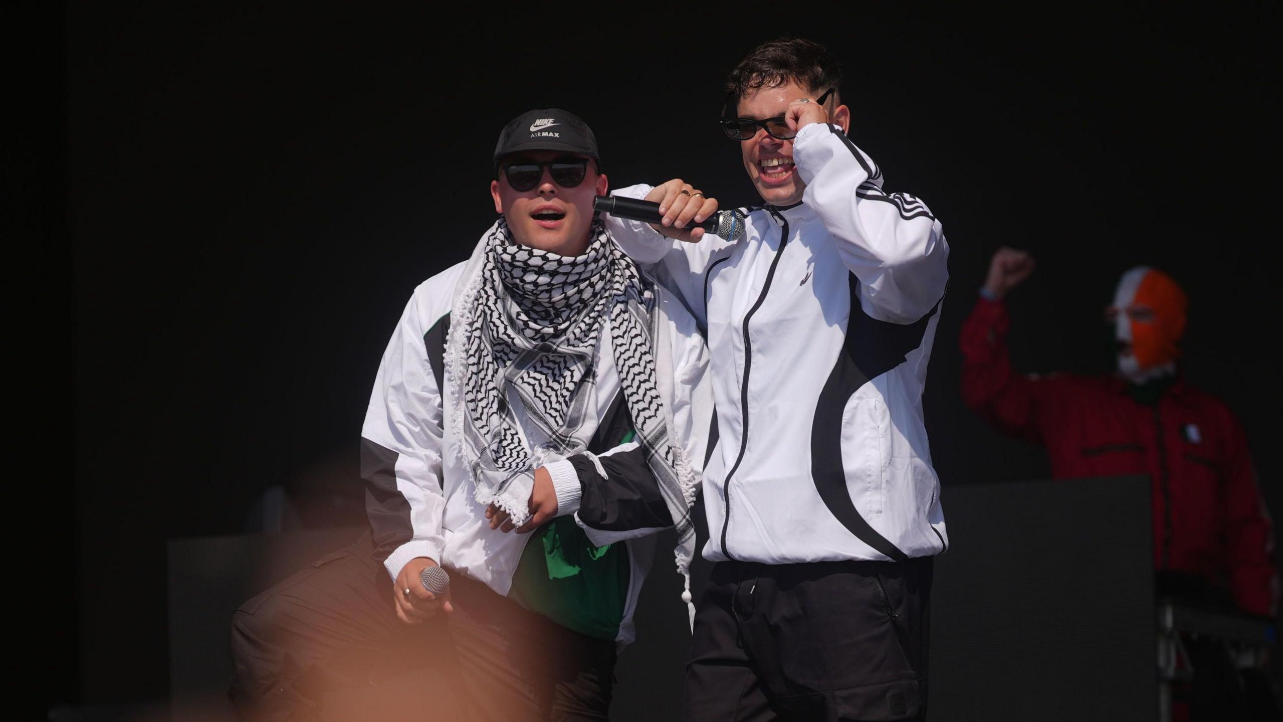 Two band members stand side by side on stage. They are both wearing white jackets and each hold a microphone. One of them has their elbow resting on his bandmate's shoulder as he appears to lift his sunglasses up. In the background, the third member can be seen holding his fist in the air. He is wearing a red jacket and a balaclava in the colours of the Irish flag.
