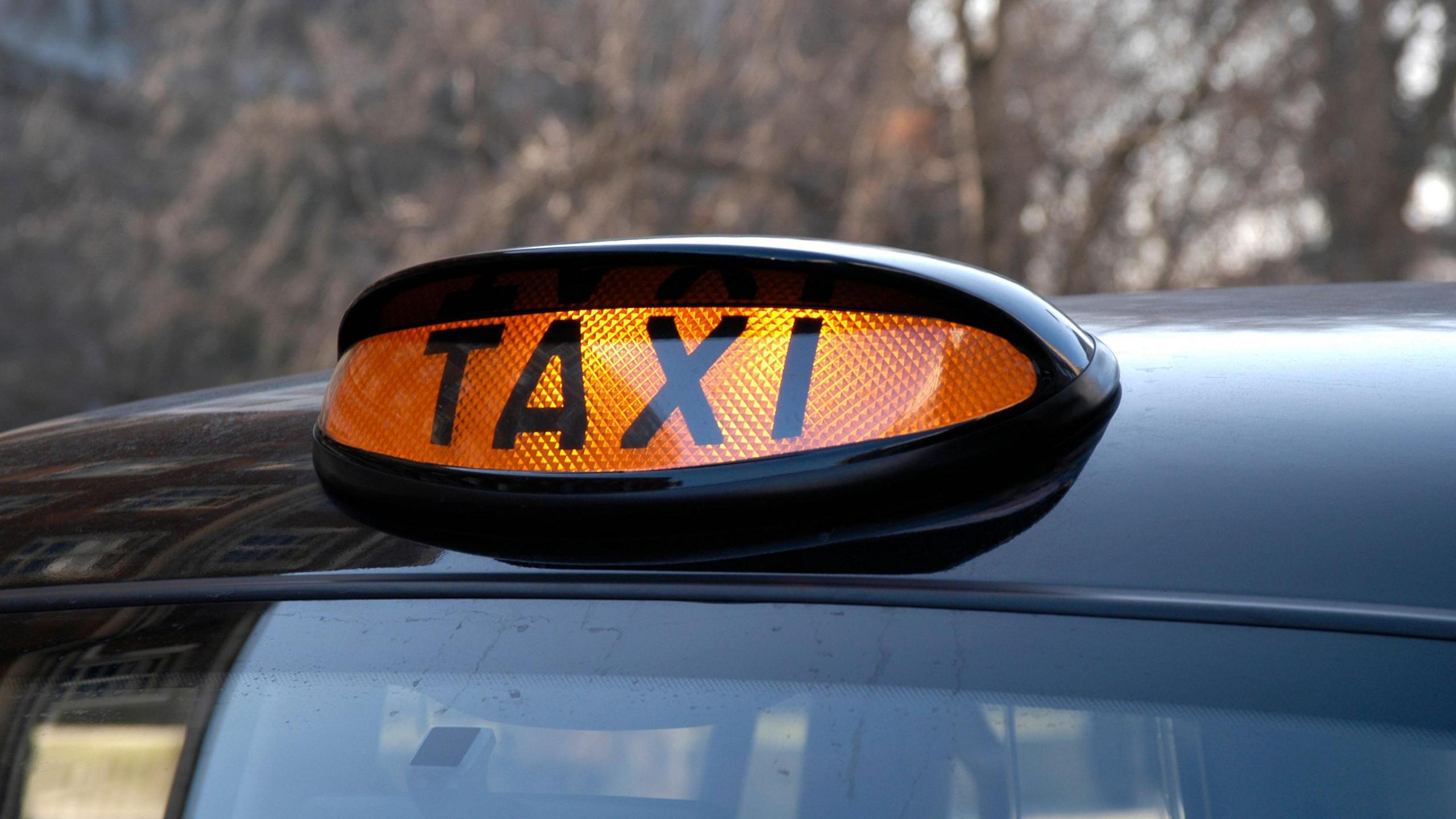 The top of a black taxi cab. The roof has an orange illuminated sign that reads "taxi" in capital letters. 