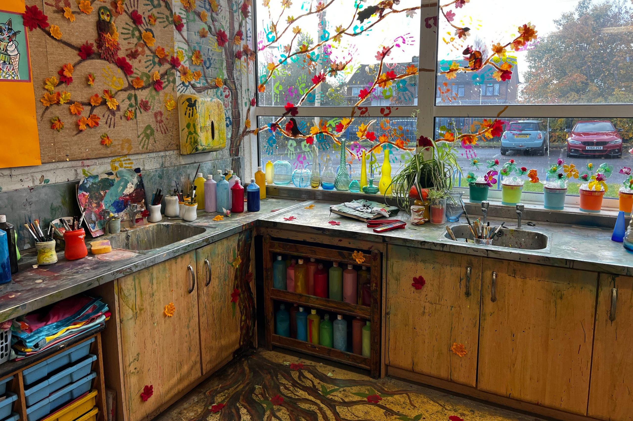 The corner of an art classroom used for filming Heartstopper. The counter tops and sinks are covered in bottles of paint and there is a mural of a tree with autumn leaves painted on the wall and along the window, its roots painted on to the floor