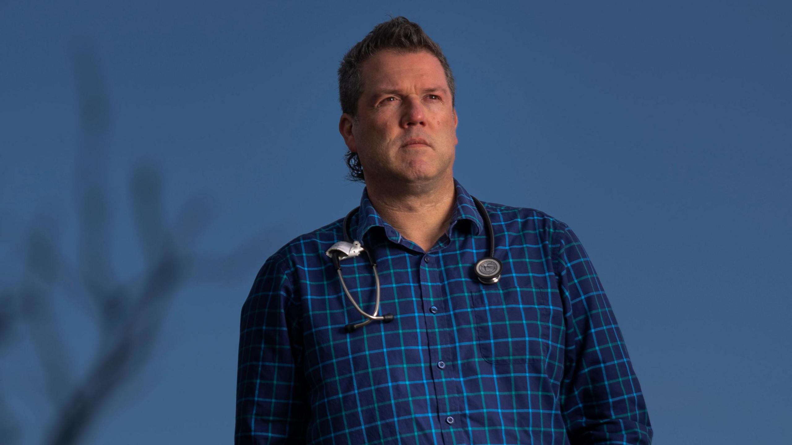 Dr Chris Webster standing outside, a bare tree in the foreground. A stethoscope is around his neck and he is wearing a blue check shirt.