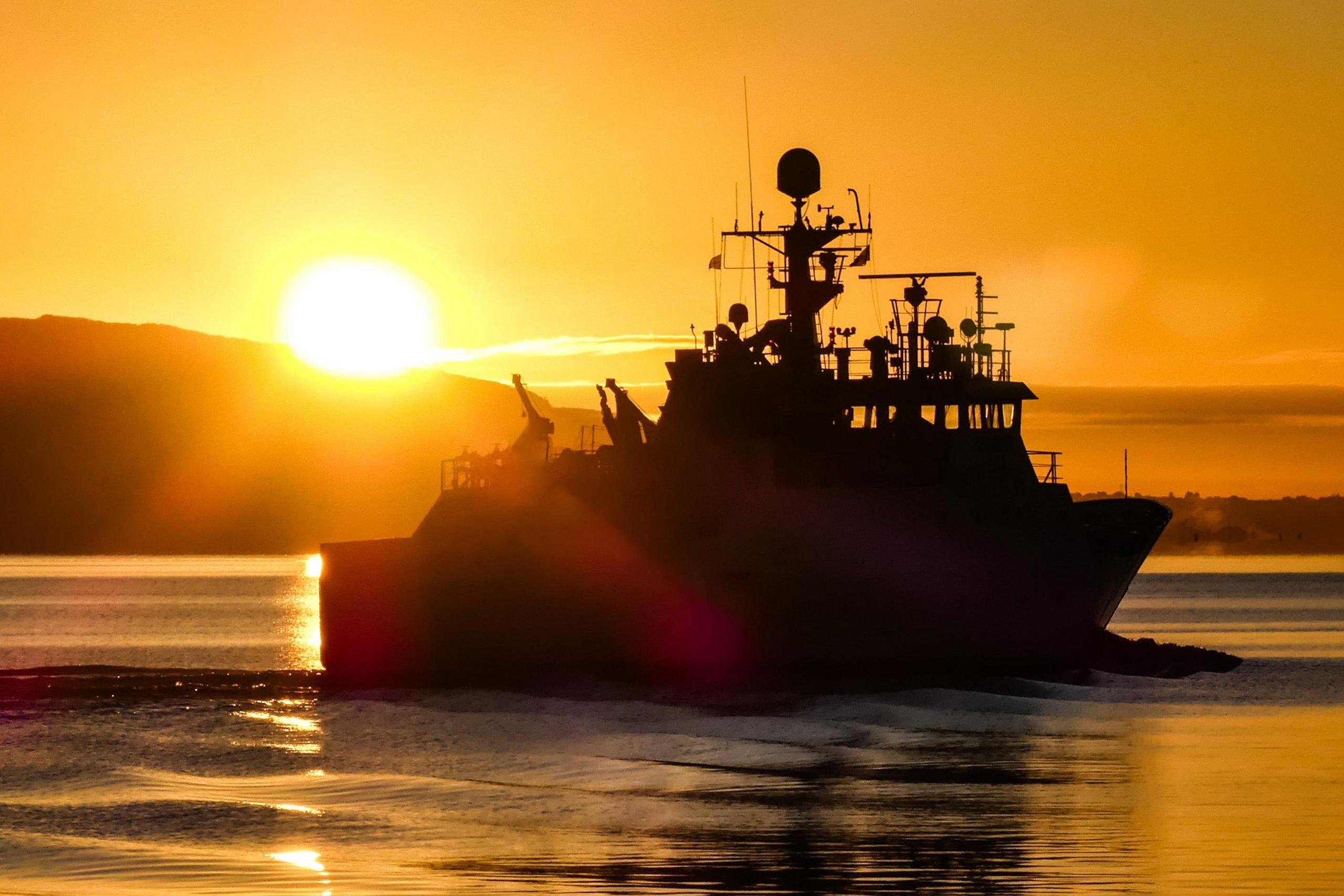 Fishery patrol vessel Minna returning on the River Clyde, with bright sun in the background

