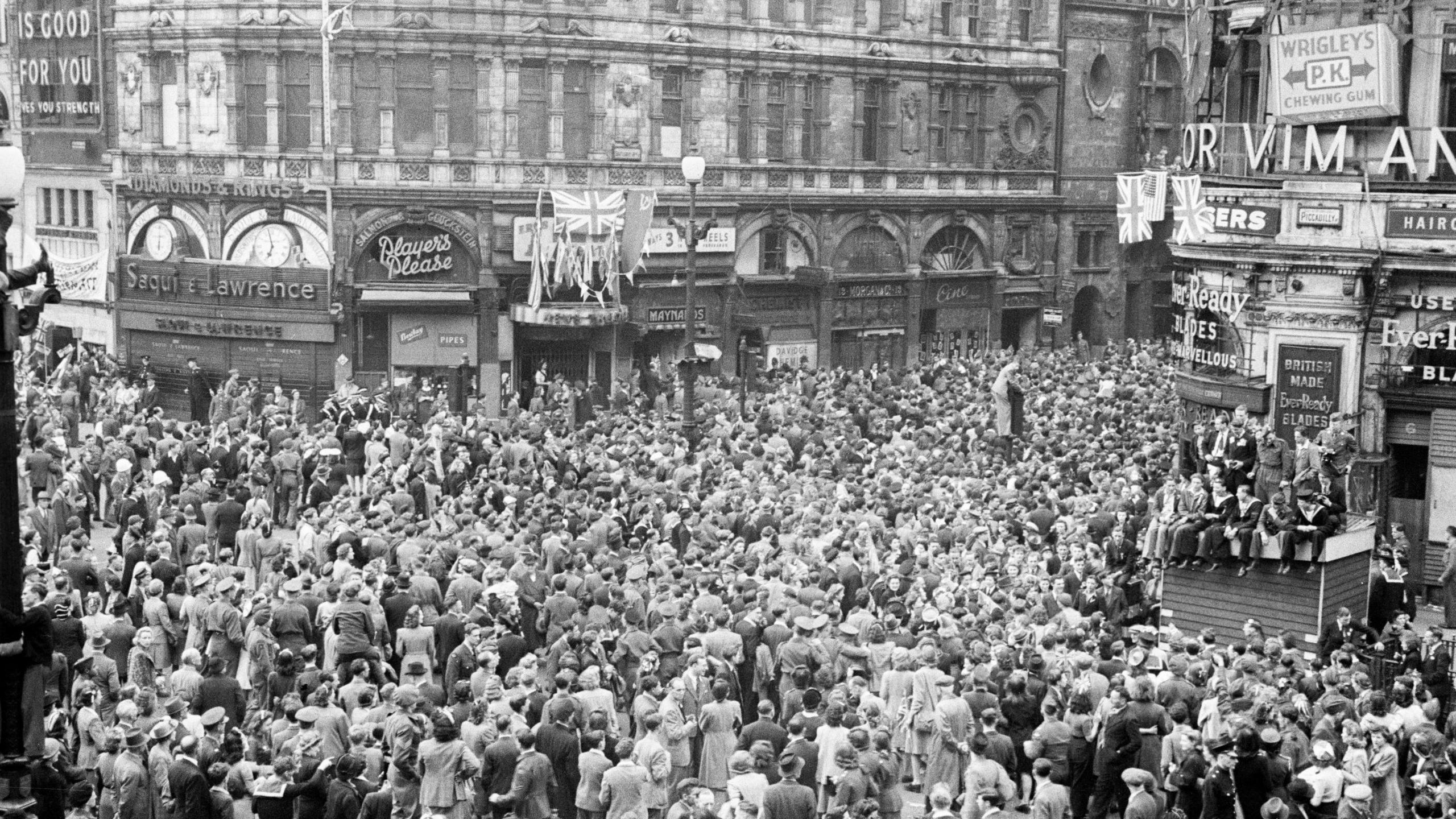 A black and white image showing hundreds of people gathered at Piccadilly Circus in London, with Union Jack flags flying from some buildings.