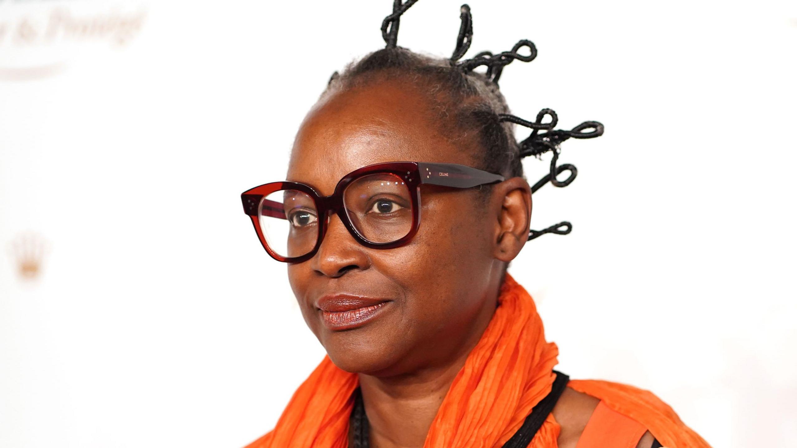 Koyo Kouoh wears thick rimmed glasses, an orange scarf and an orange top.