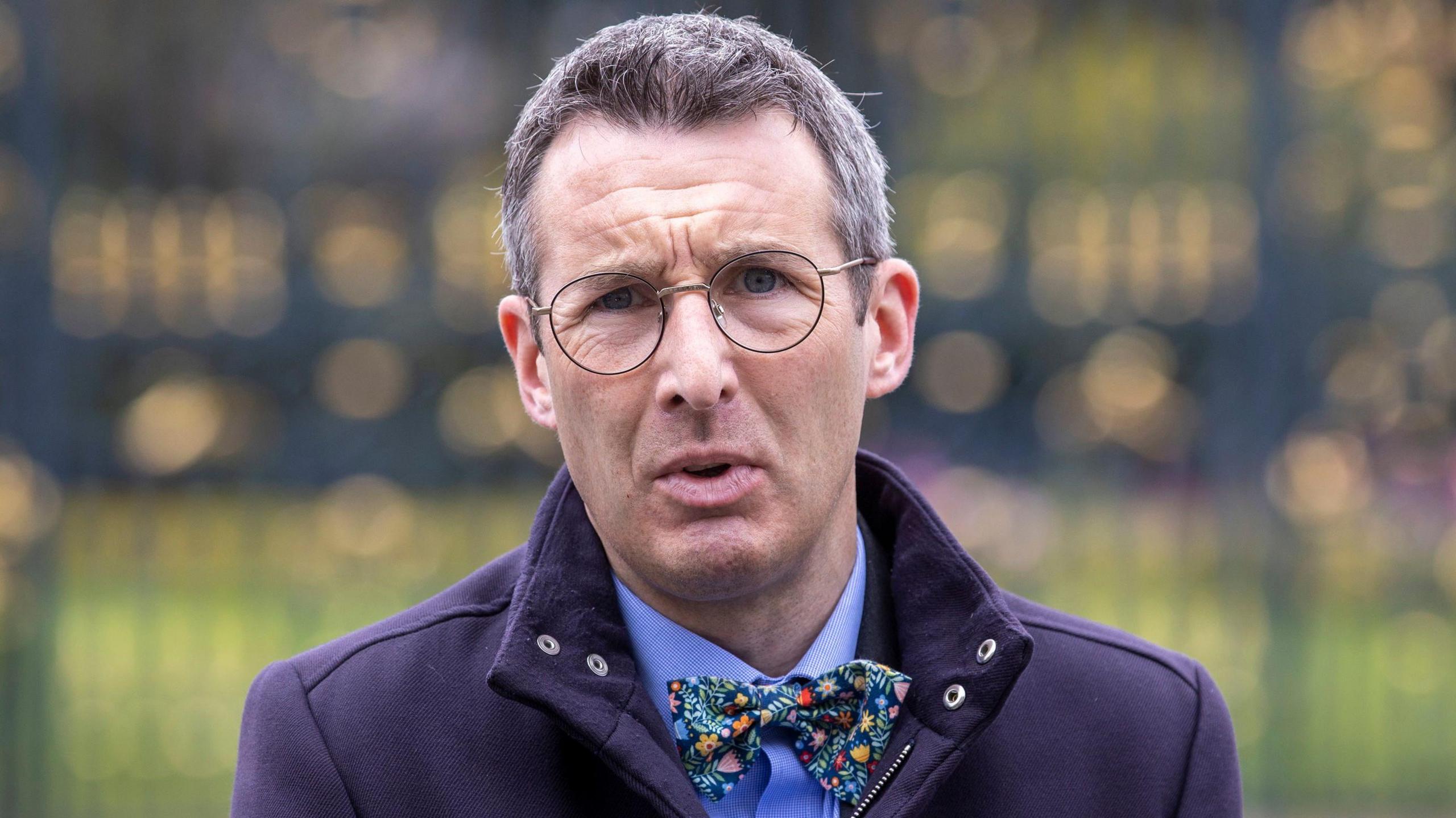 Andrew Muir wears glasses. He has a navy coat on and a blue shirt with a floral bow tie.