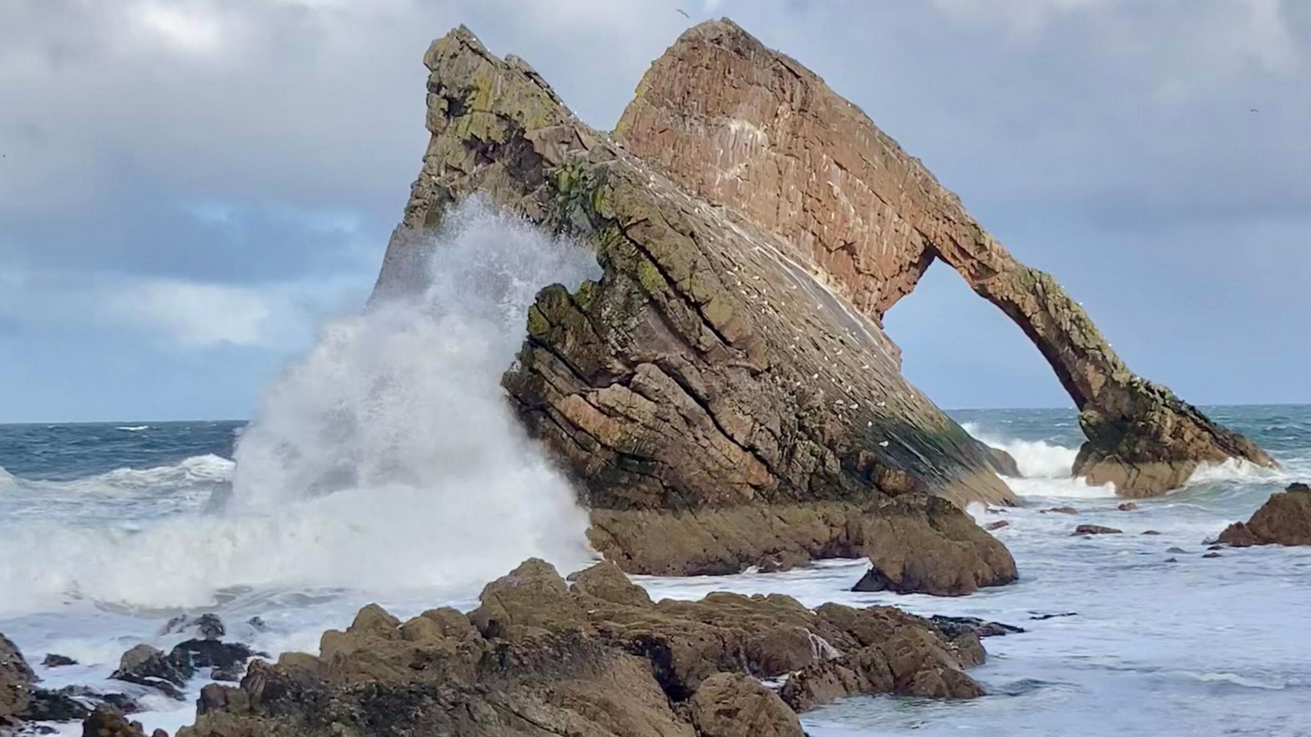 Two large rocks in the sea. One in the background has a large arch-shaped hole on its right side. A thin strip of rock extends into the sea, which is white and foamy. In the foreground, a rock with a large white wave crashing against it sits just behind a small outcrop of rocks in the water.