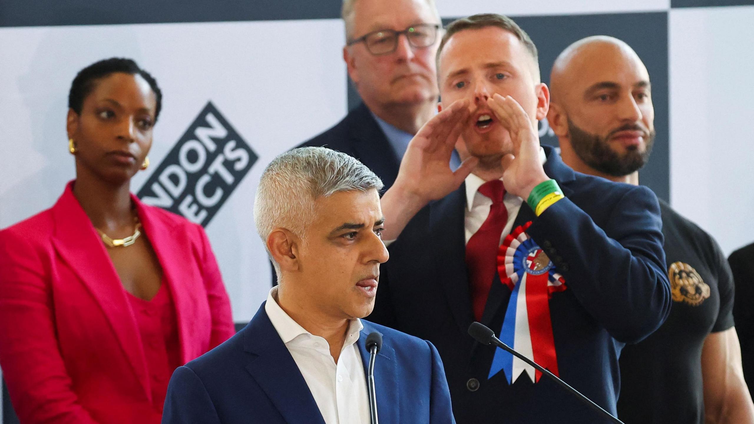 Nick Scanlon, a white man with a red, white and blue rosette representing Britain First, raises his hands to his mouth while shouting at London mayor Sadiq Khan. 