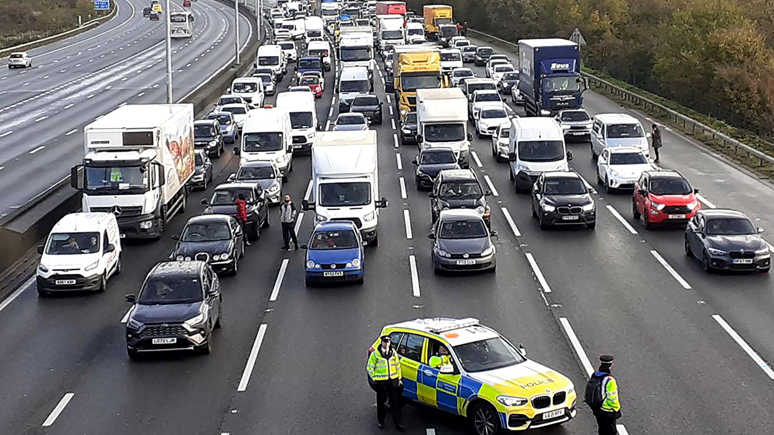 Traffic stopped on M25, lined up like in a car park, with some people standing outside their cars, and a police car with three officers