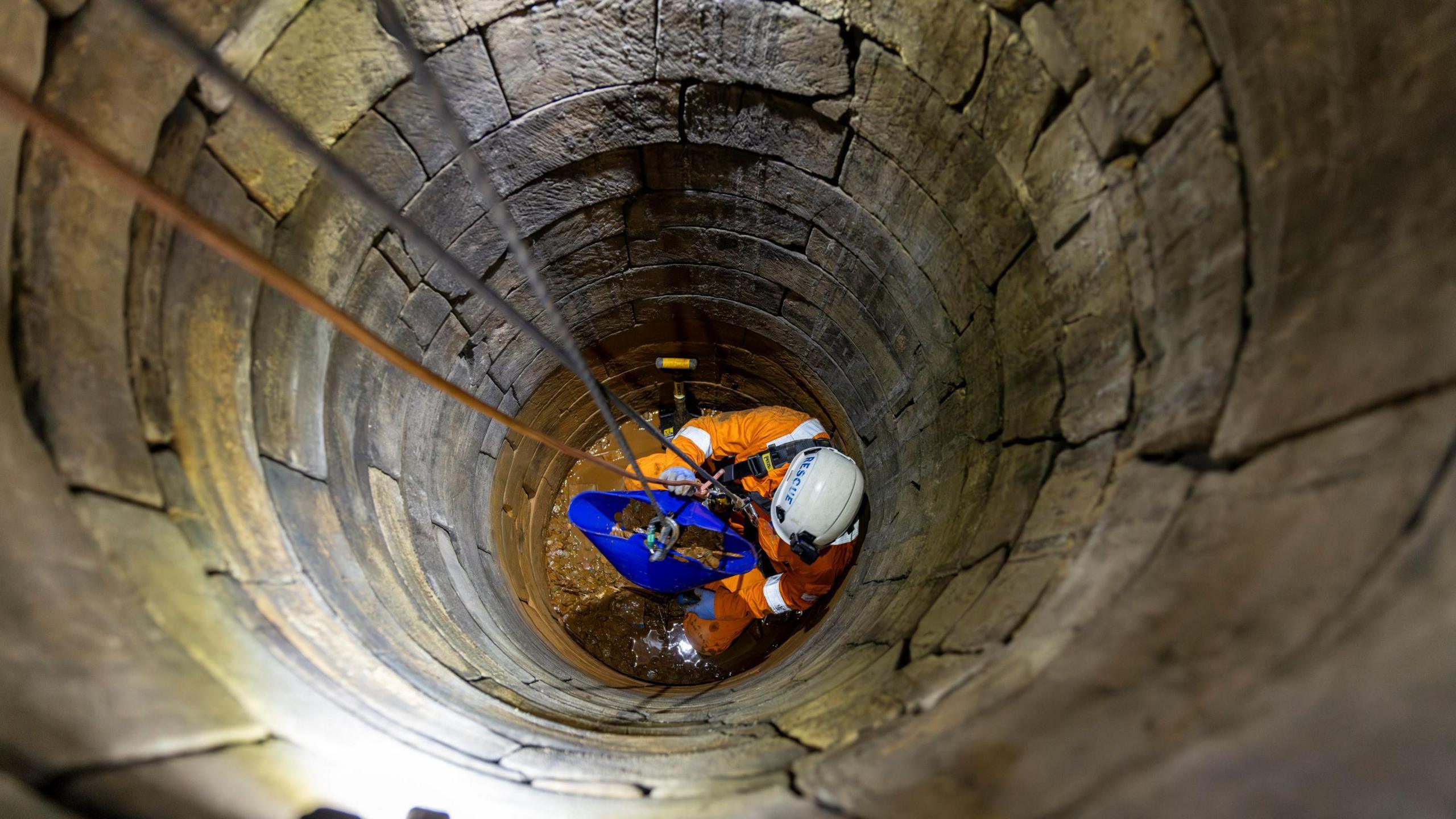 A man dressed in hi-vis work gear and a helmet is lowered to the bottom of a stone well. He has filled a blue bucket with copper coins which he is about to send up on a rope.