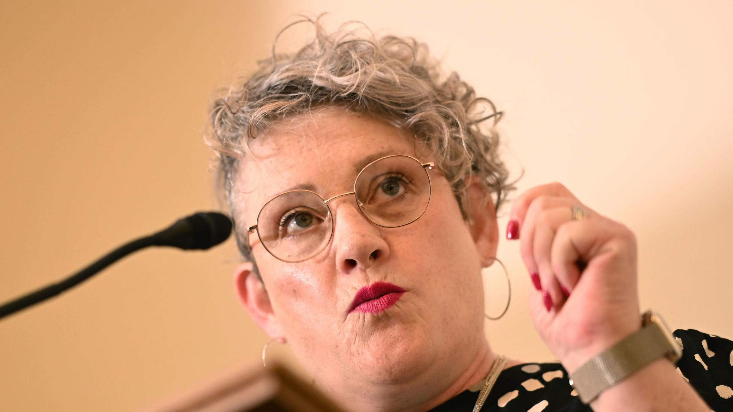 A close up of Ashley Dalton as she speaks from a lectern into a microphone. She has short, curly grey hair and is wearing glasses and large hoop earrings. She is wearing a black and white spotted top and her nails are painted red.