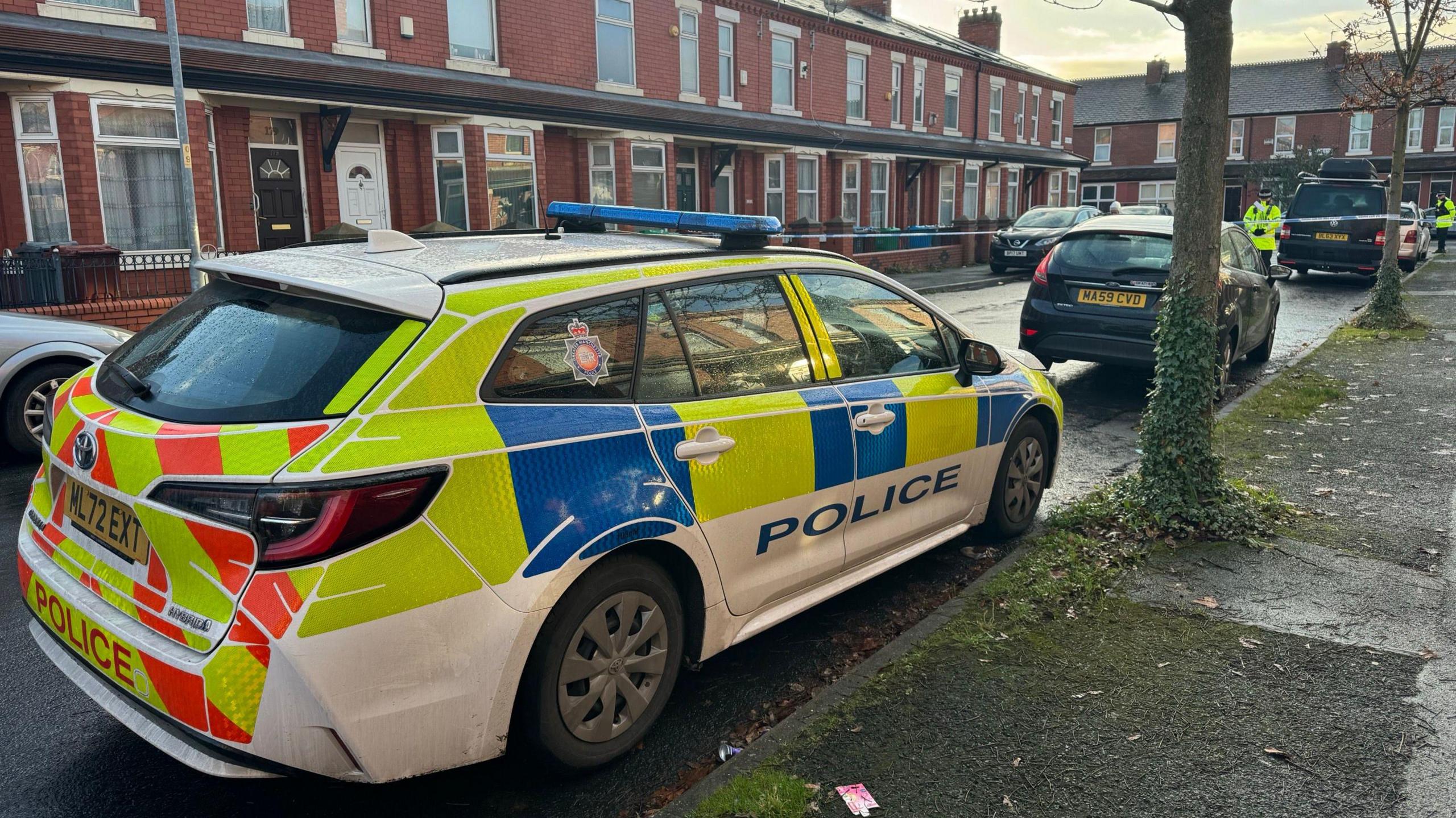 A marked police car is parked just outside a taped-off cordon on Great Southern Street in Moss Side. There is a row of redbrick terraced houses on one side of the road.