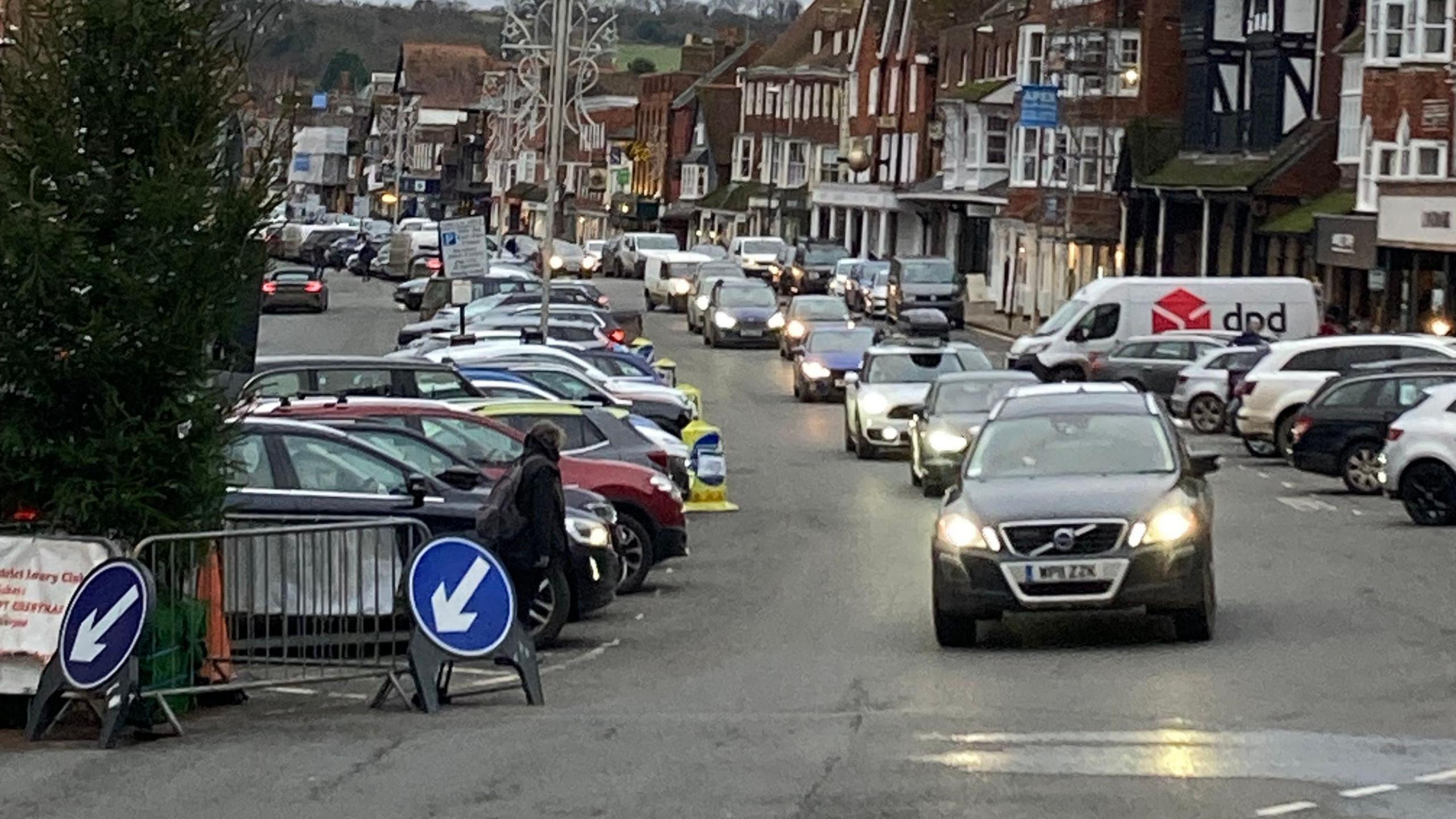 Looking down the High Street with parking down the middle, a long stream of cars on the right with historic buildings next to them.