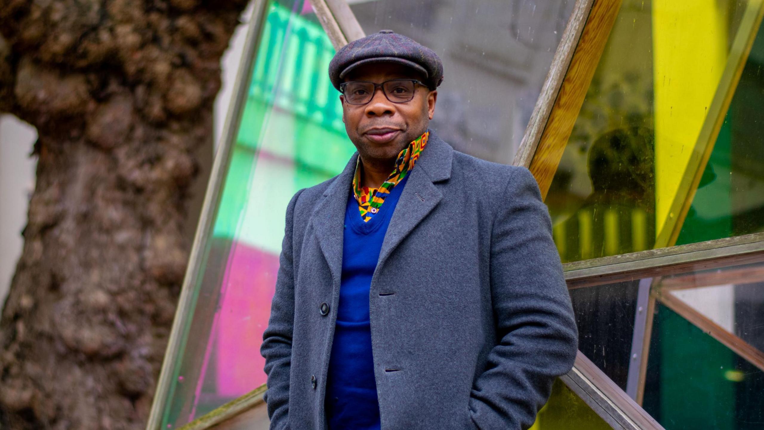 Dr Edson Burton stands facing the camera outside a University of Bristol building. He is wearing a grey woollen-style jacket with a blue jumper, a brightly-coloured yellow shirt and a grey cloth cap.