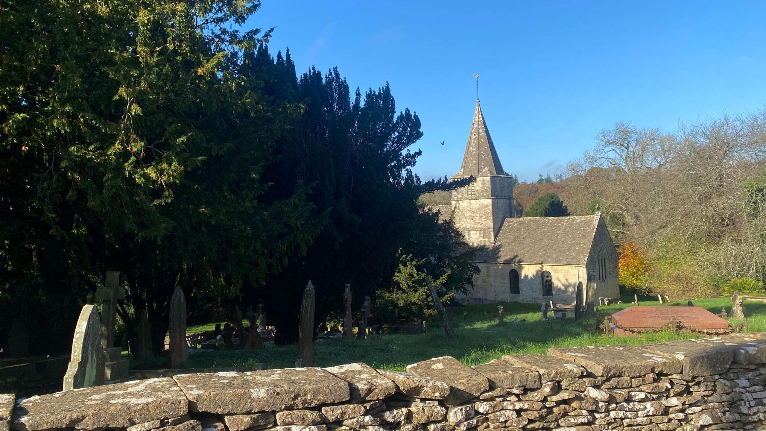 The exterior of St Kenelm's in Sapperton. A small church with a pointed cone-shaped spire is visible in the background, with the graveyard in the foreground. 