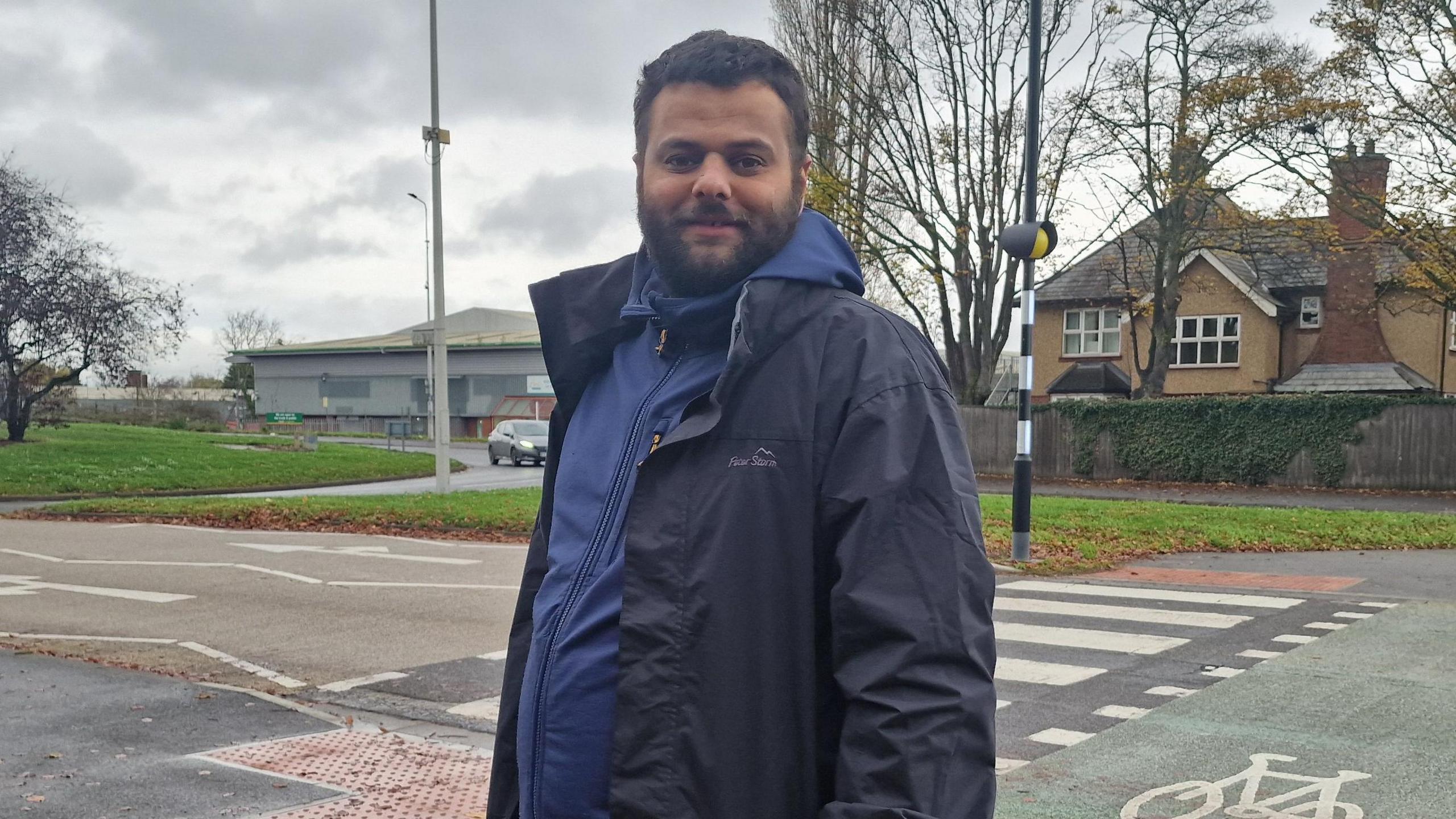 A man, Samir Own, with short dark hair and a beard, wearing a blue fleece and blue jacket, standing in front of a zebra crossing. There is a car, a residential building, a commercial building and a roundabout with a tree on it in the background.
