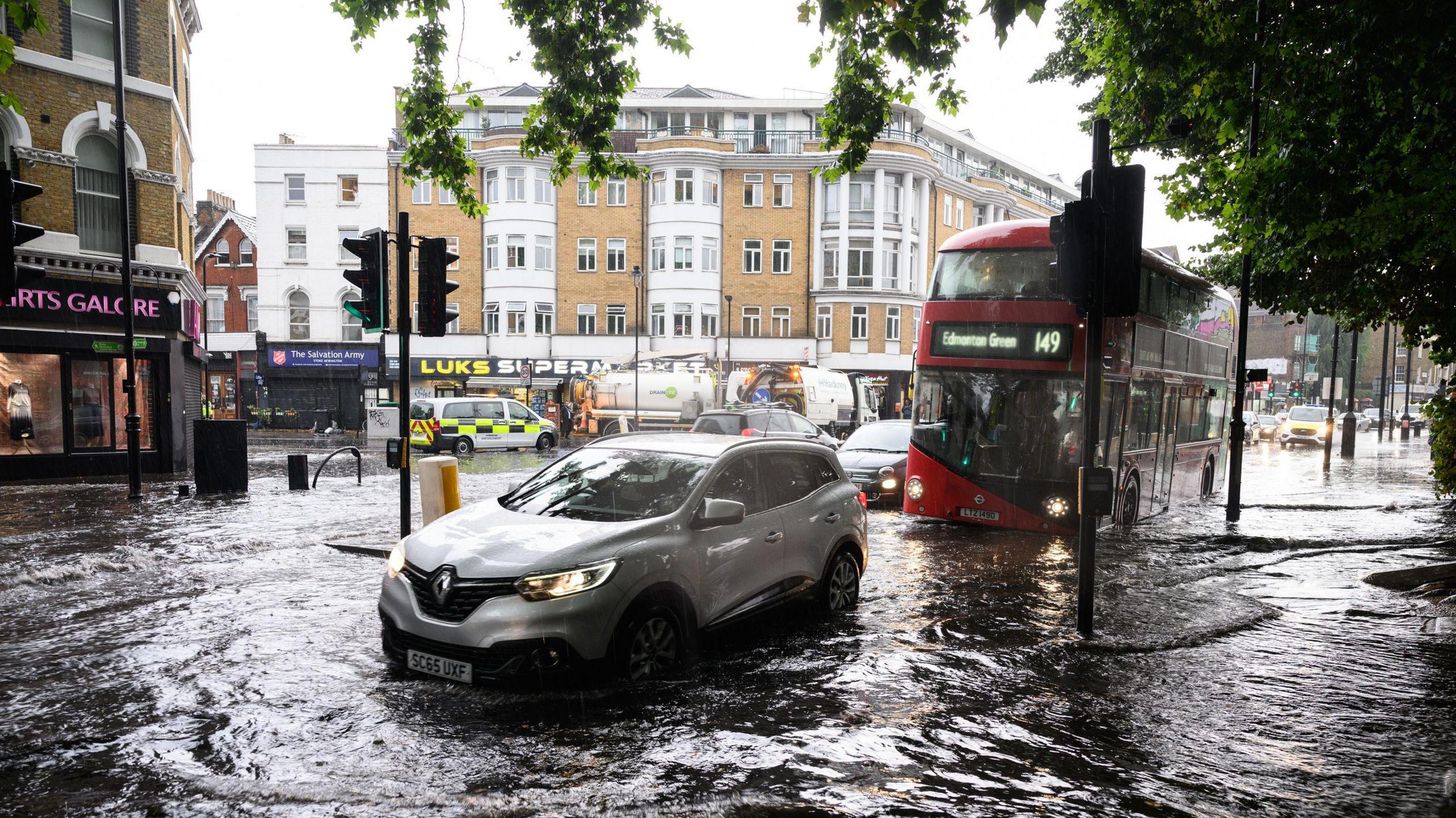 A flooded London street, with a silver car and red London bus driving through the water at a crossing. There are flats and shops visible in the background and a tree hangs over the street, visible in the top right of the picture