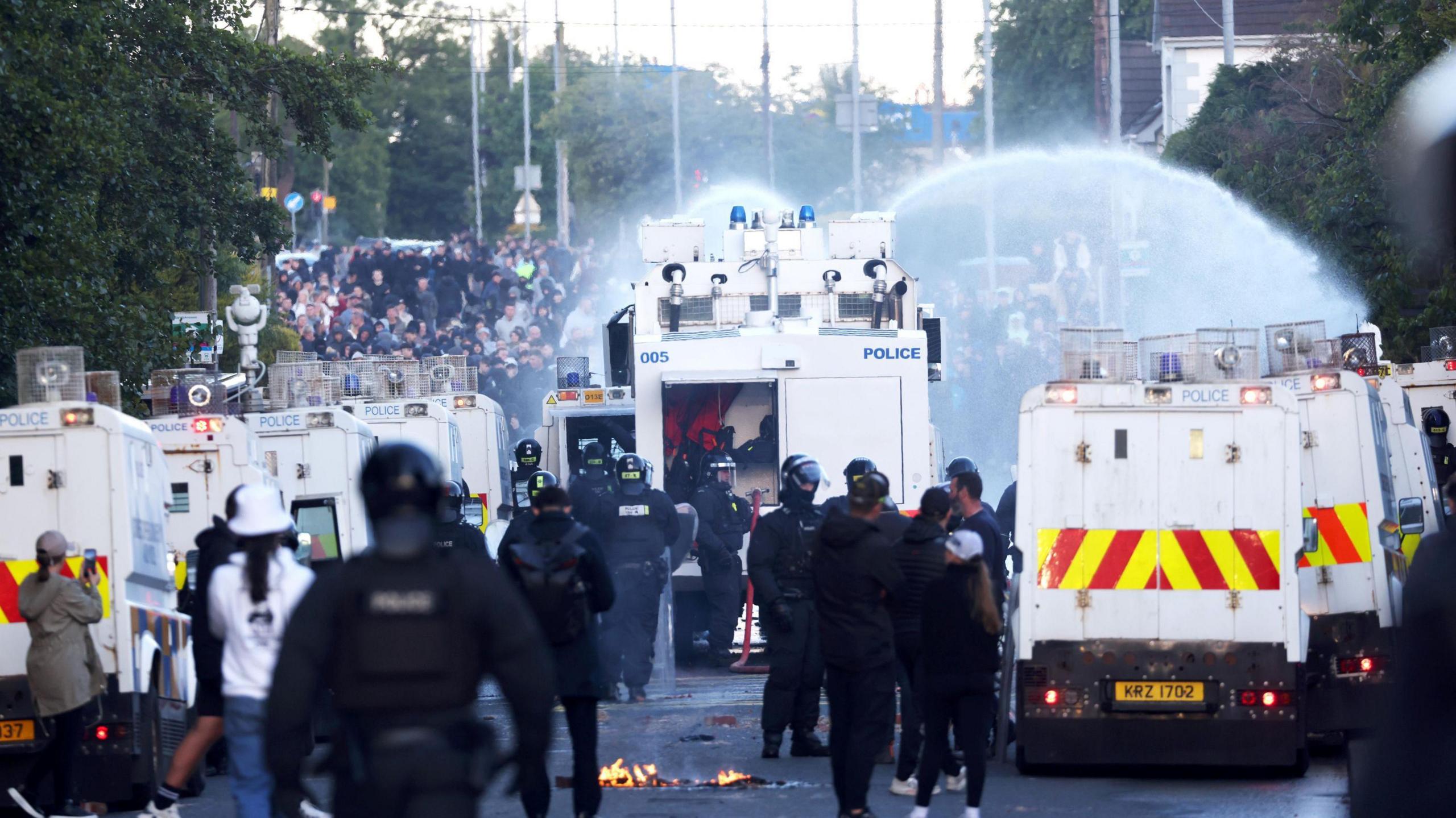 A number of police vans and police in riot car in the middle of a road with the remains of a fire beside one the vans. There is a water cannon near the front spraying a large crowd of people.