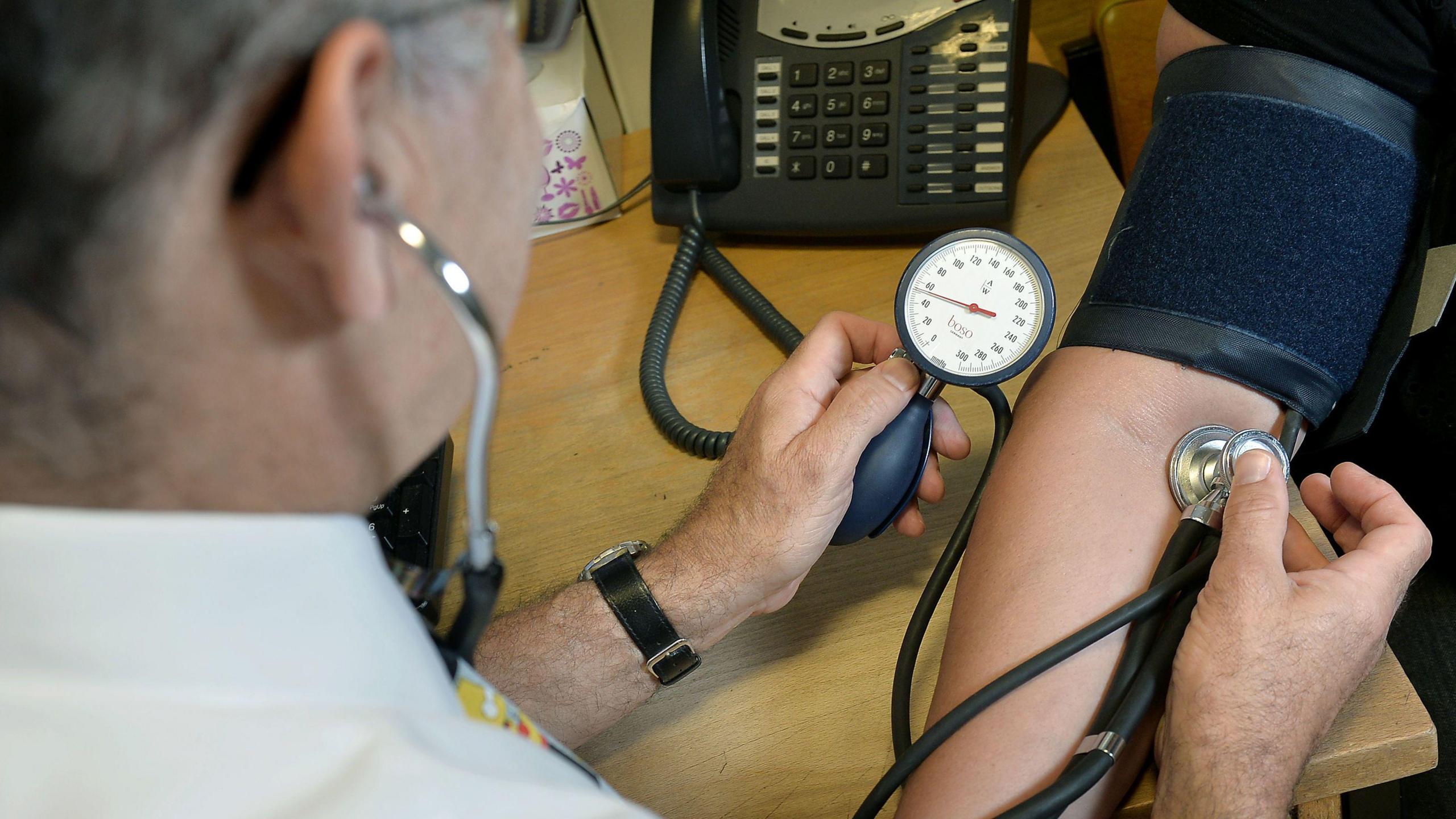A stock image of a man in a white coat using blood pressure equipment on a patient's arm. The equipment includes a measuring clock, a round silver stethoscope and a blue arm wrap.