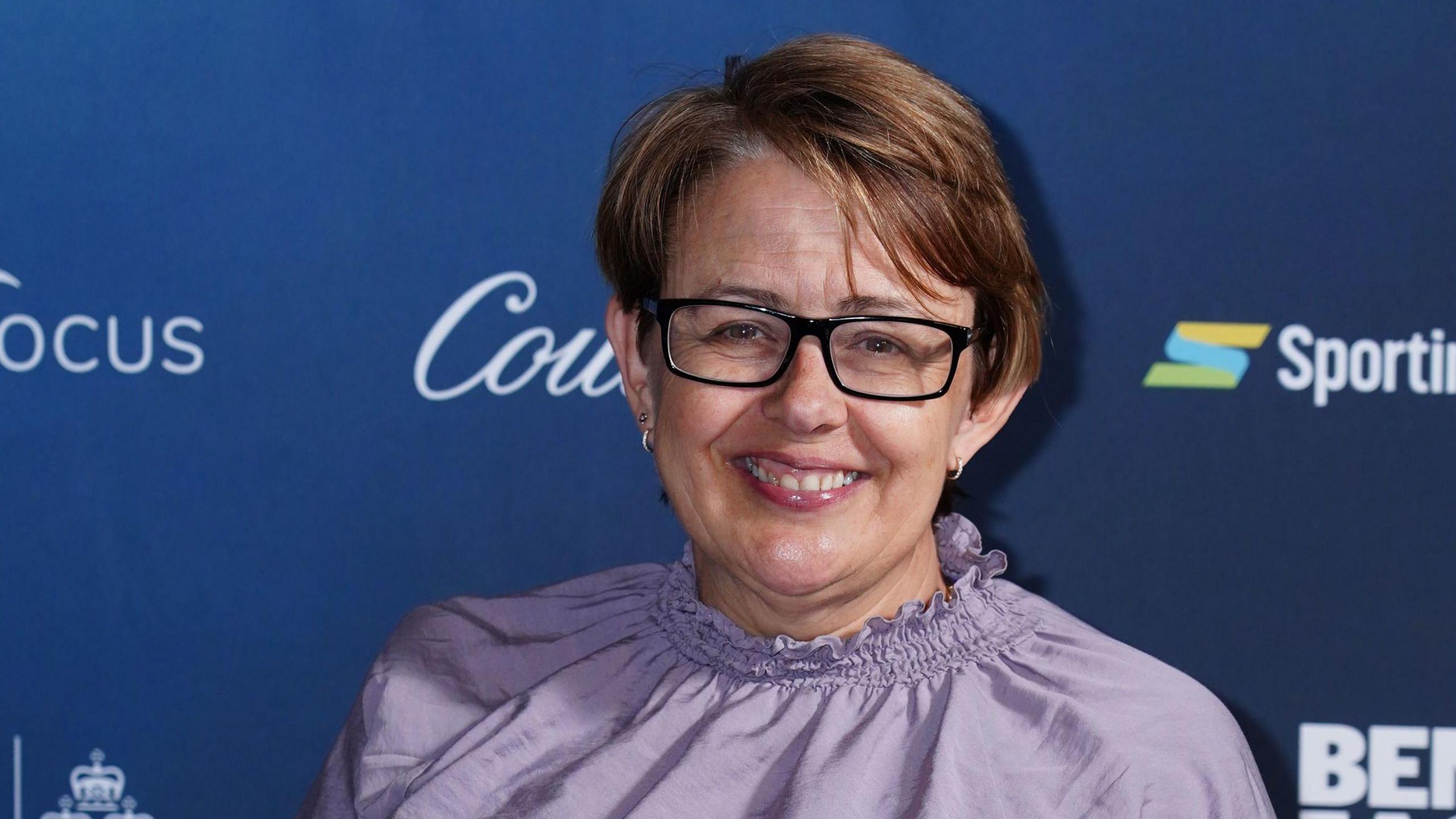 Tanni Grey-Thompson is smiling for the camera on a red carpet. She has short brown hair and black glasses.