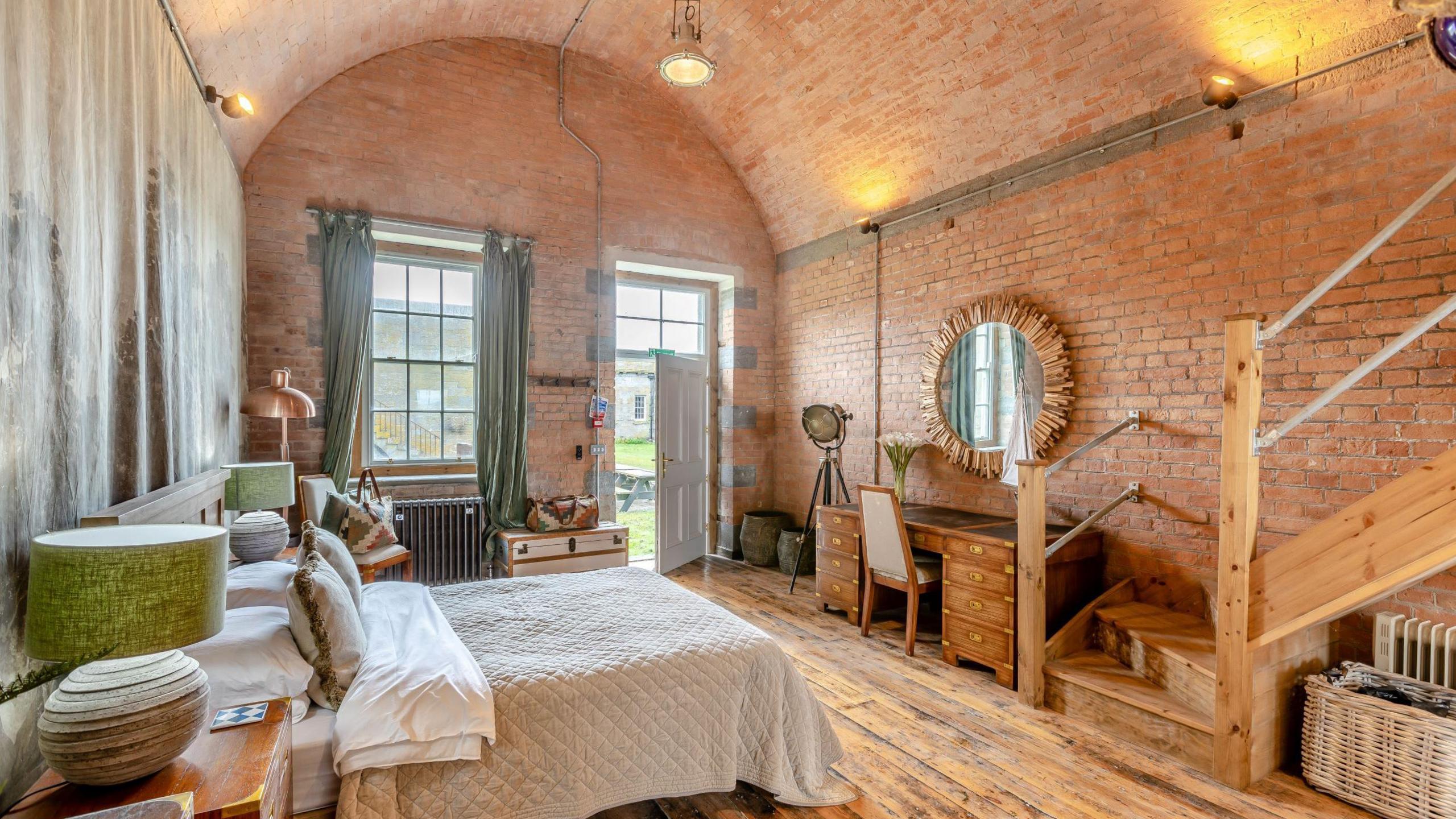 A bedroom with exposed red brick walls. A bed sits against one wall, with a wooden dresser opposite and a set of wooden steps leading out of view. A set of windows and a door leading outdoors can be seen on the furthest wall. 