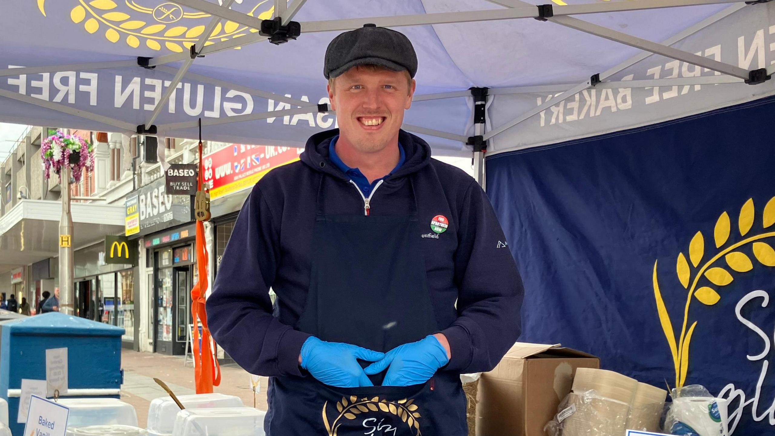 Tom standing in a market stall, under a gazebo. He is wearing a flat cap and has blue plastic gloves on his hands. He is in Southend high street.