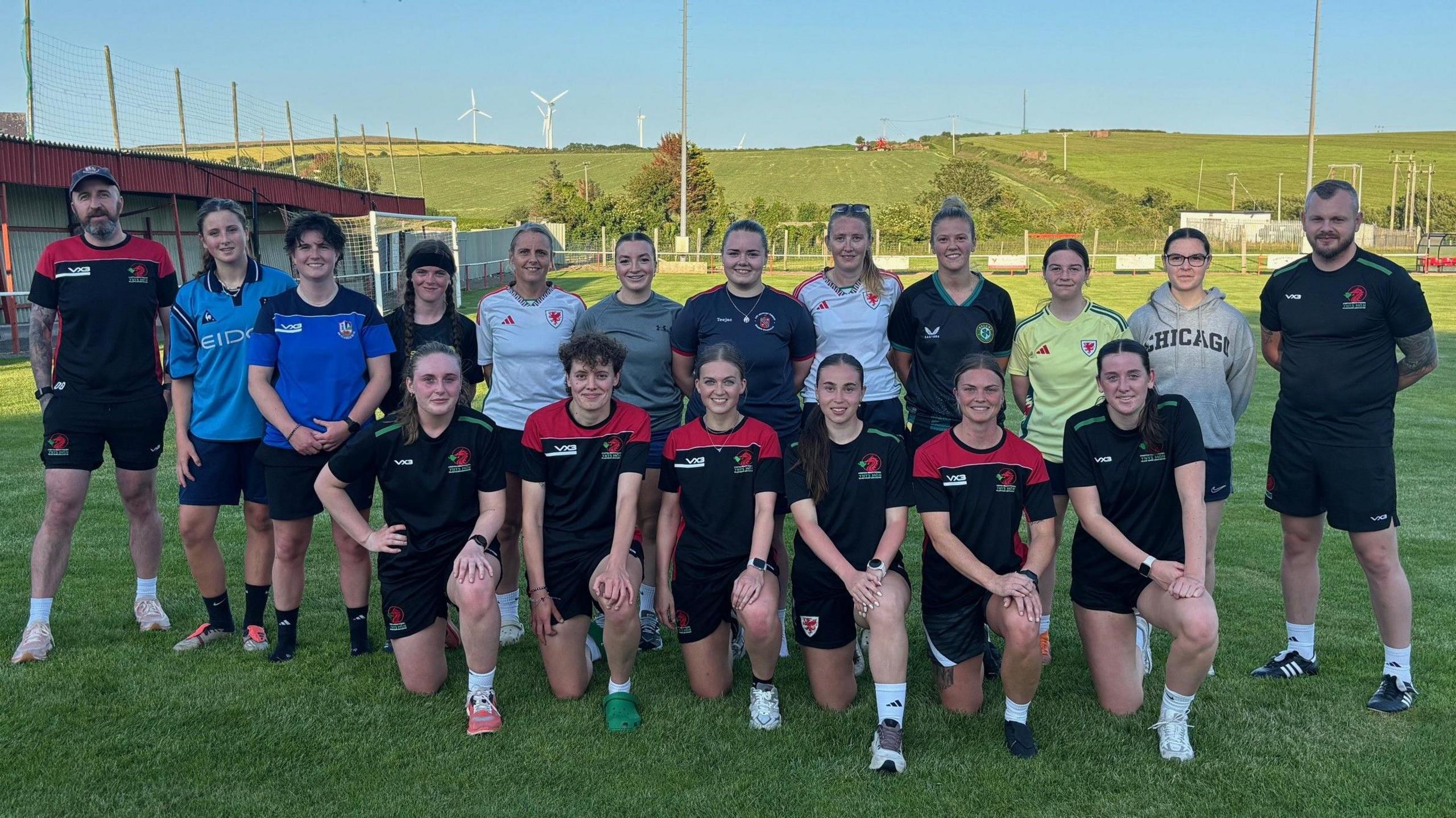 Tîm pêl-droed merched Môn yn sefyll ymysg ei gilydd gyda'u hyfforddwyr ar gae chwarae Cemaes. Mae 18 ohonyn nhw gyda'i gilydd mewn dwy res yn wynebu'r camera. Mae'r haul yn machlud, ac maent yn gwenu ar y camera. Mae rhai ohonynt yn gwisgo crysau chwarae lliwgar a'r gweddill yn gwisgo crys pêl-droed coch a du.