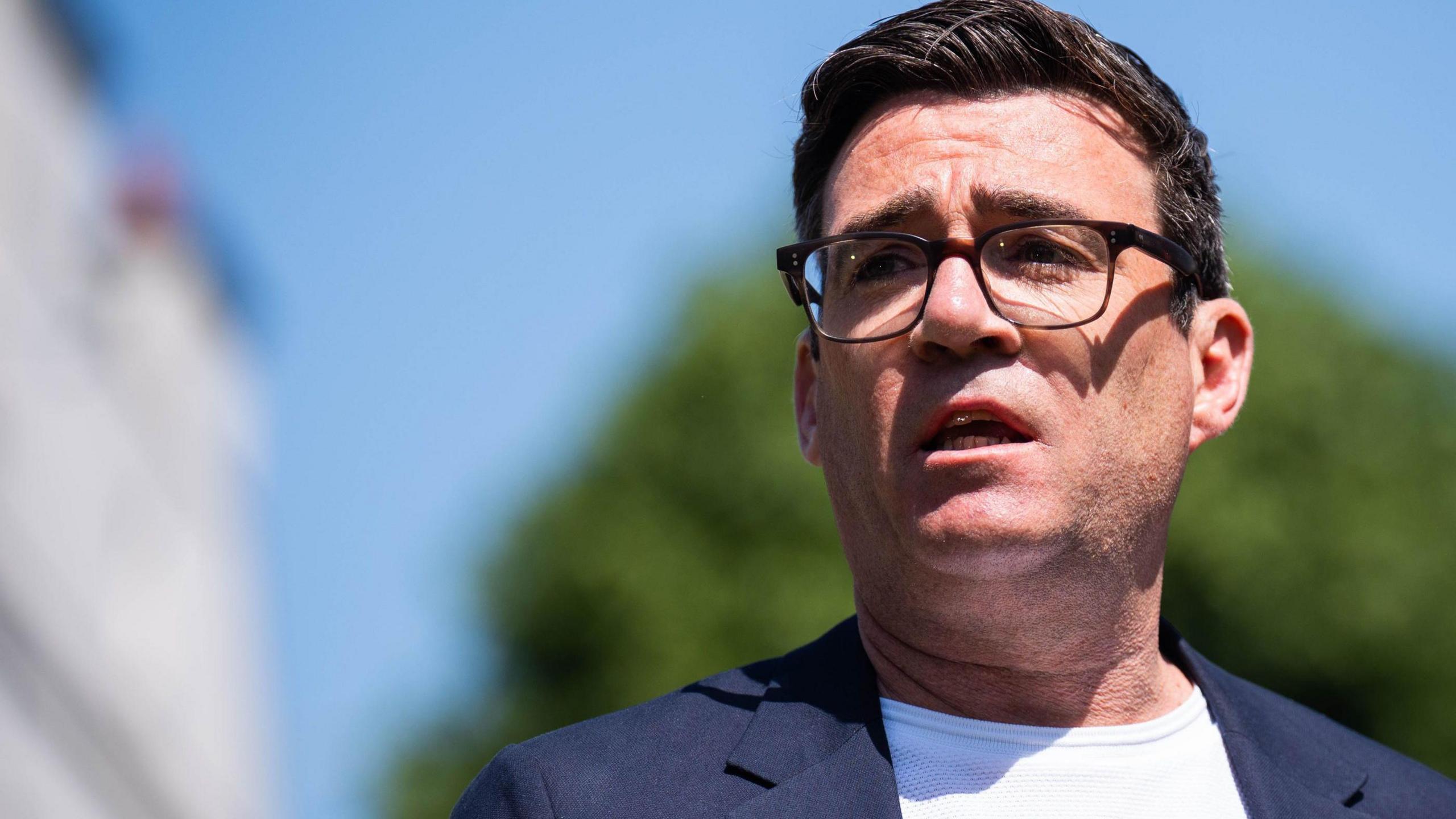 Mayor of Greater Manchester Andy Burnham wearing a white t-shirt and blue suit jacket and glasses. 