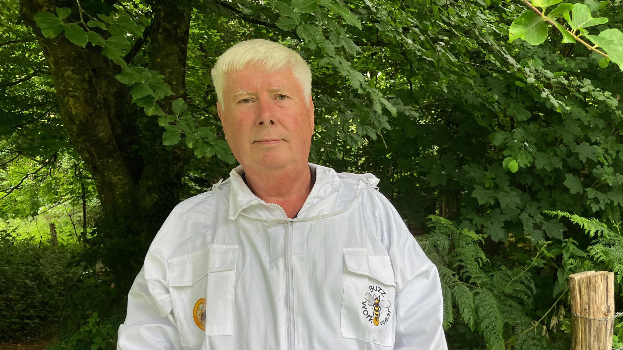 A grey haired man, wearing a white protective beekeeping suit, stood in front of a wooded area