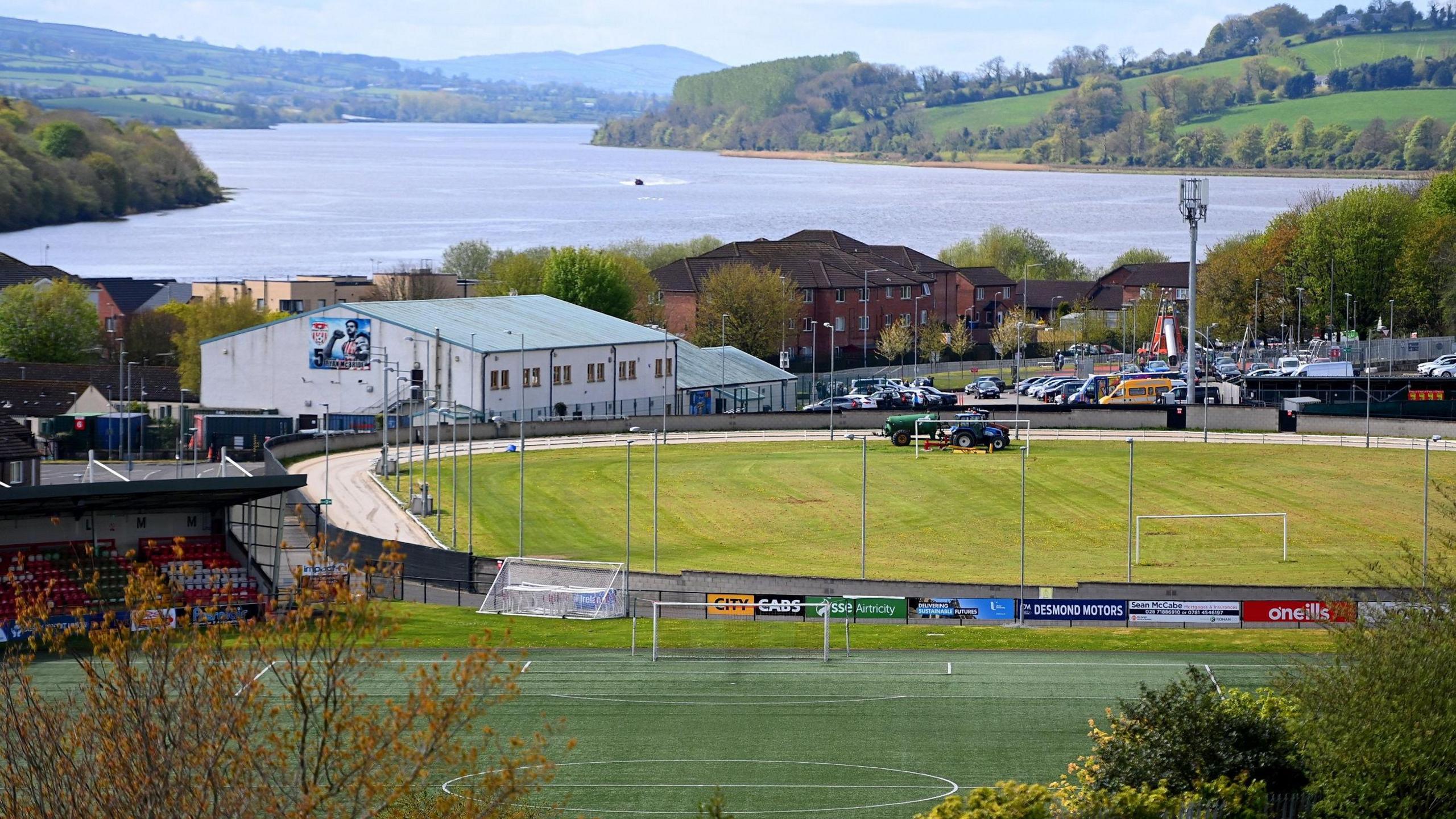 The greyhound racing track in Lononderry, with the brandywell soccer stadium in the foreground and the river Foyle in the background. A tractor can be seen in the middle of the track, the footballs ground's stand is visible to the left, while a car park is to the right, with a number of vehicles parked in it.