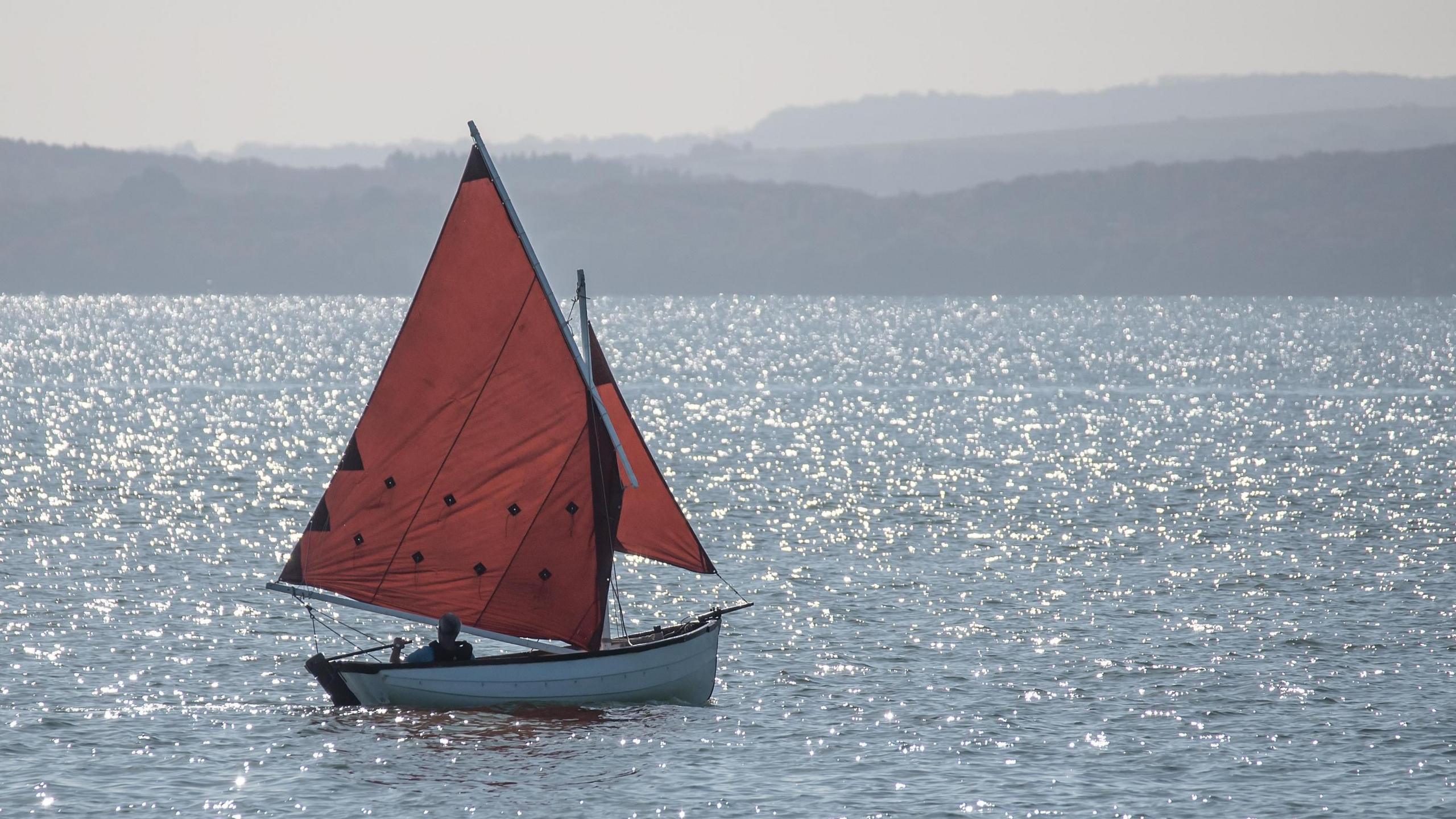 A small white dingy with red sails bobs across a shimmering sea in the bright sunlight. A person can be seen in silhouette at the tiller.