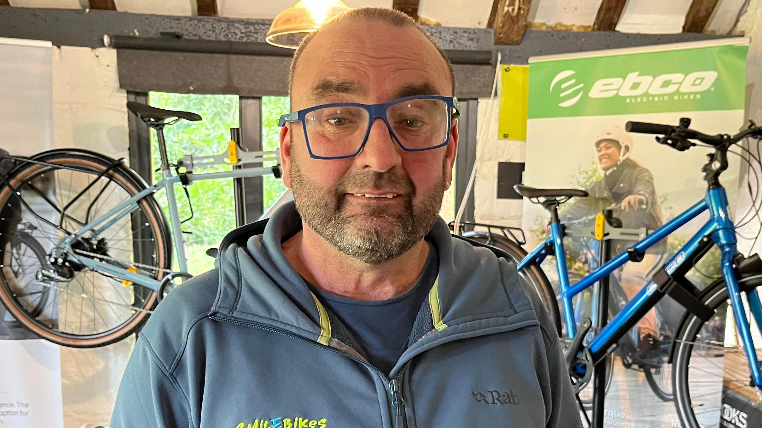 Tim Gregory, a man who is looking at the camera and smiling and is wearing a blue pair of glasses. He is wearing a blue hoodie with the logo of his business on in yellow writing. 