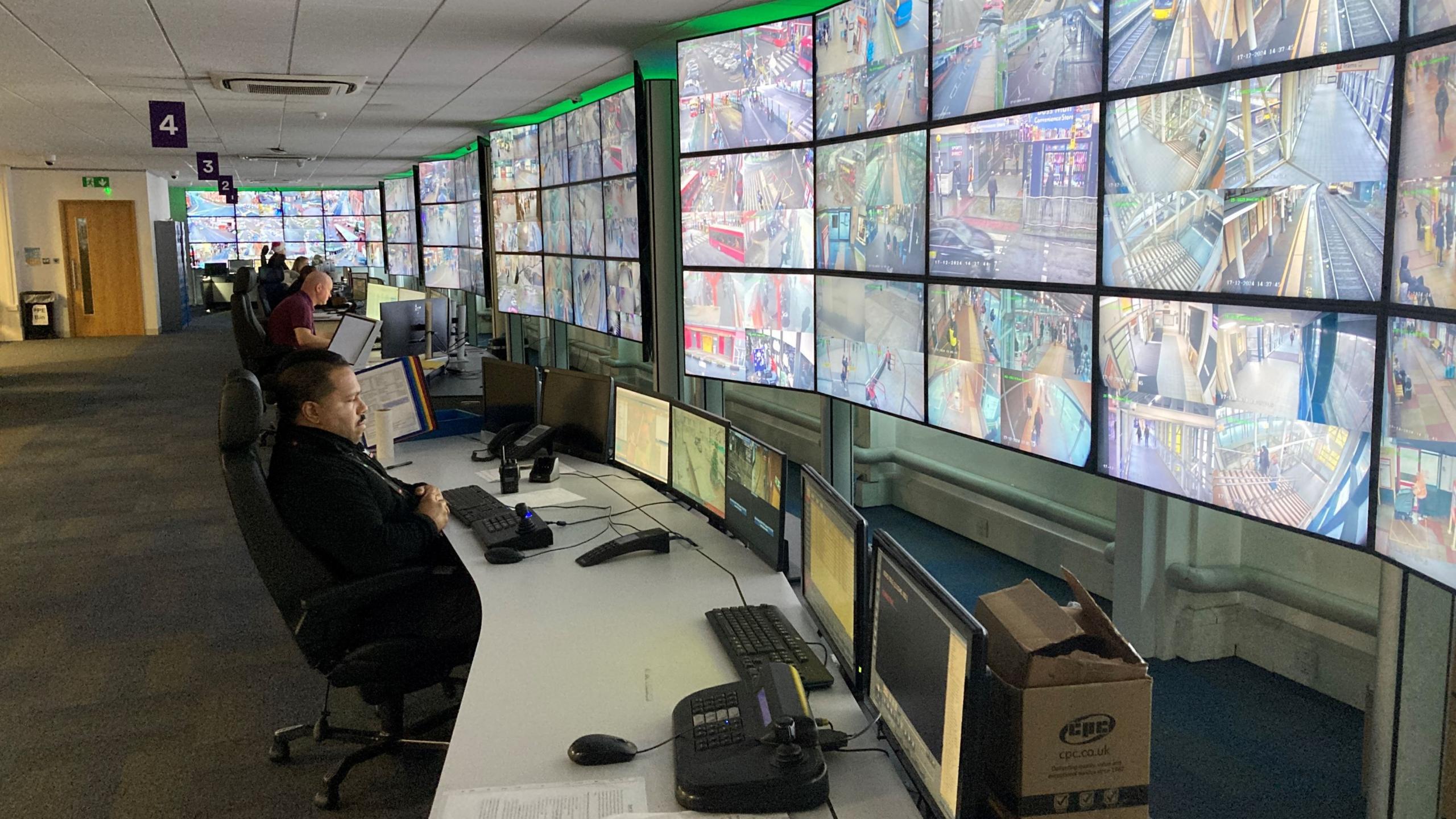 A line of workers are seated and look at many screens in front of them. The screens show different areas and some rail tracks and crossings can be seen on the screens closest to the camera. The office also has small computer screens with some mousepads and two landline phones are also in shot.